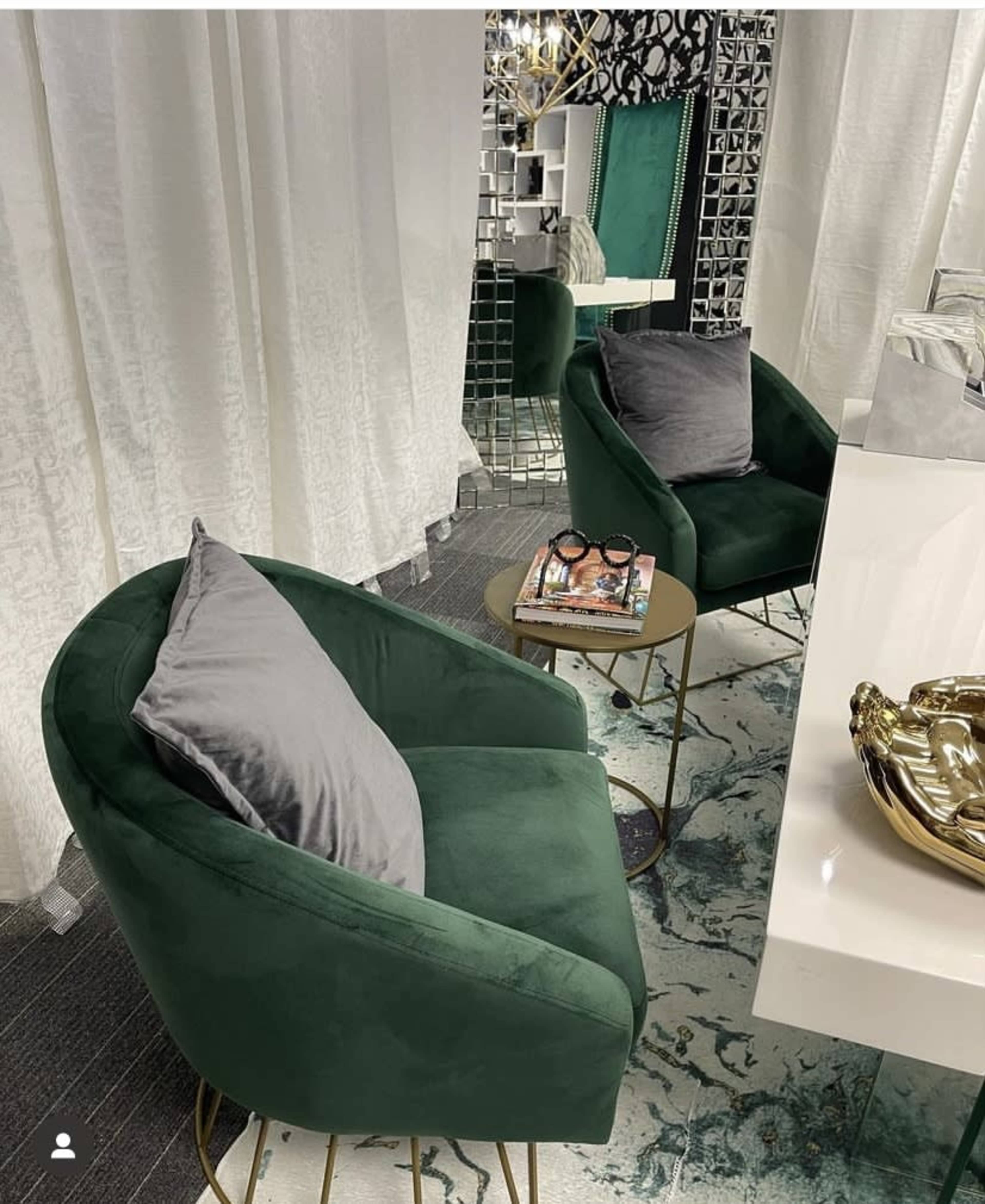 Two green velvet chairs with gray pillows positioned beside a small gold side table, which holds magazines, in a room featuring patterned walls and a mirrored accent.