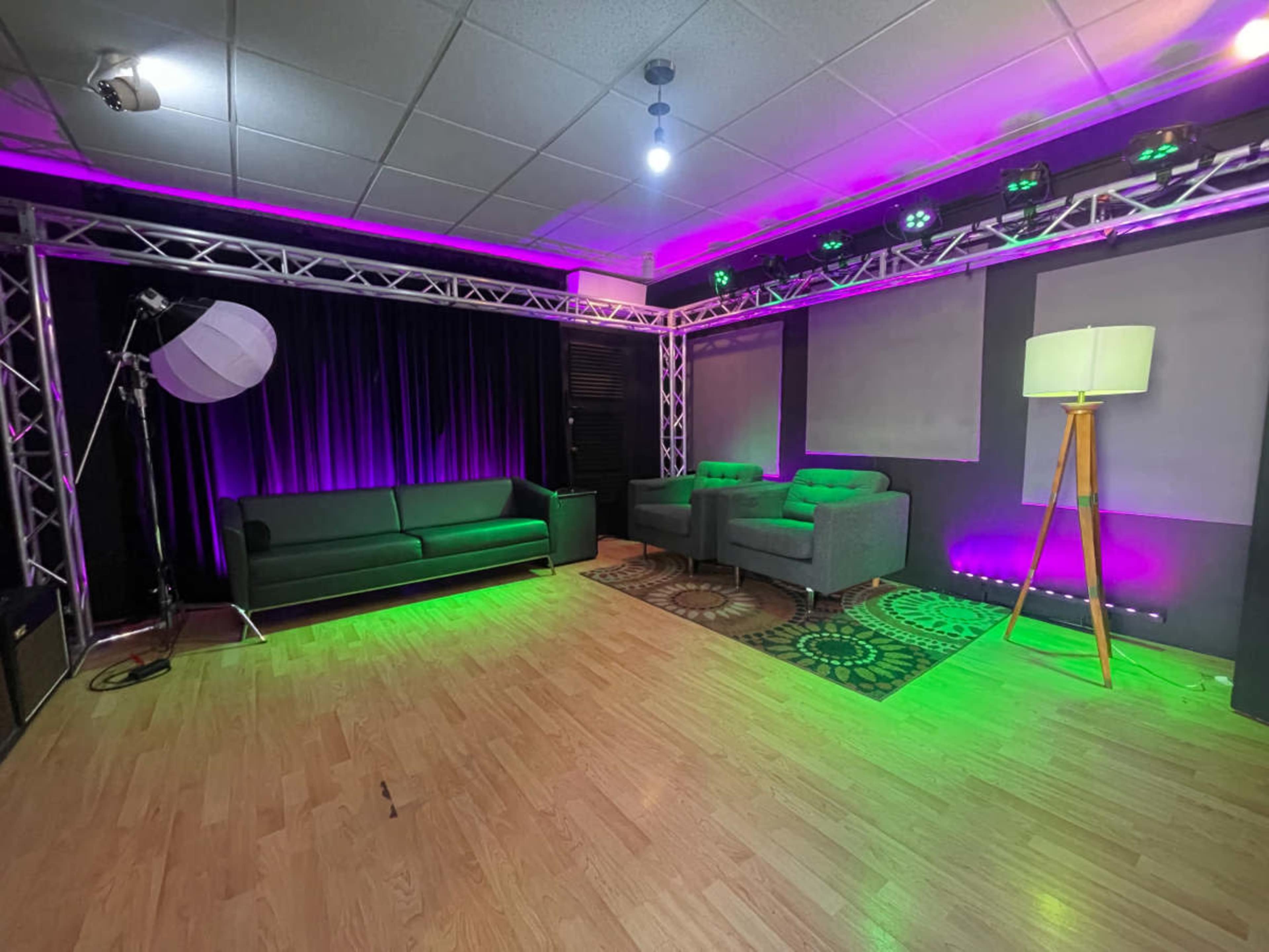 The image shows a modern studio space with a green couch, two gray armchairs, and colorful lighting along the walls and ceiling.