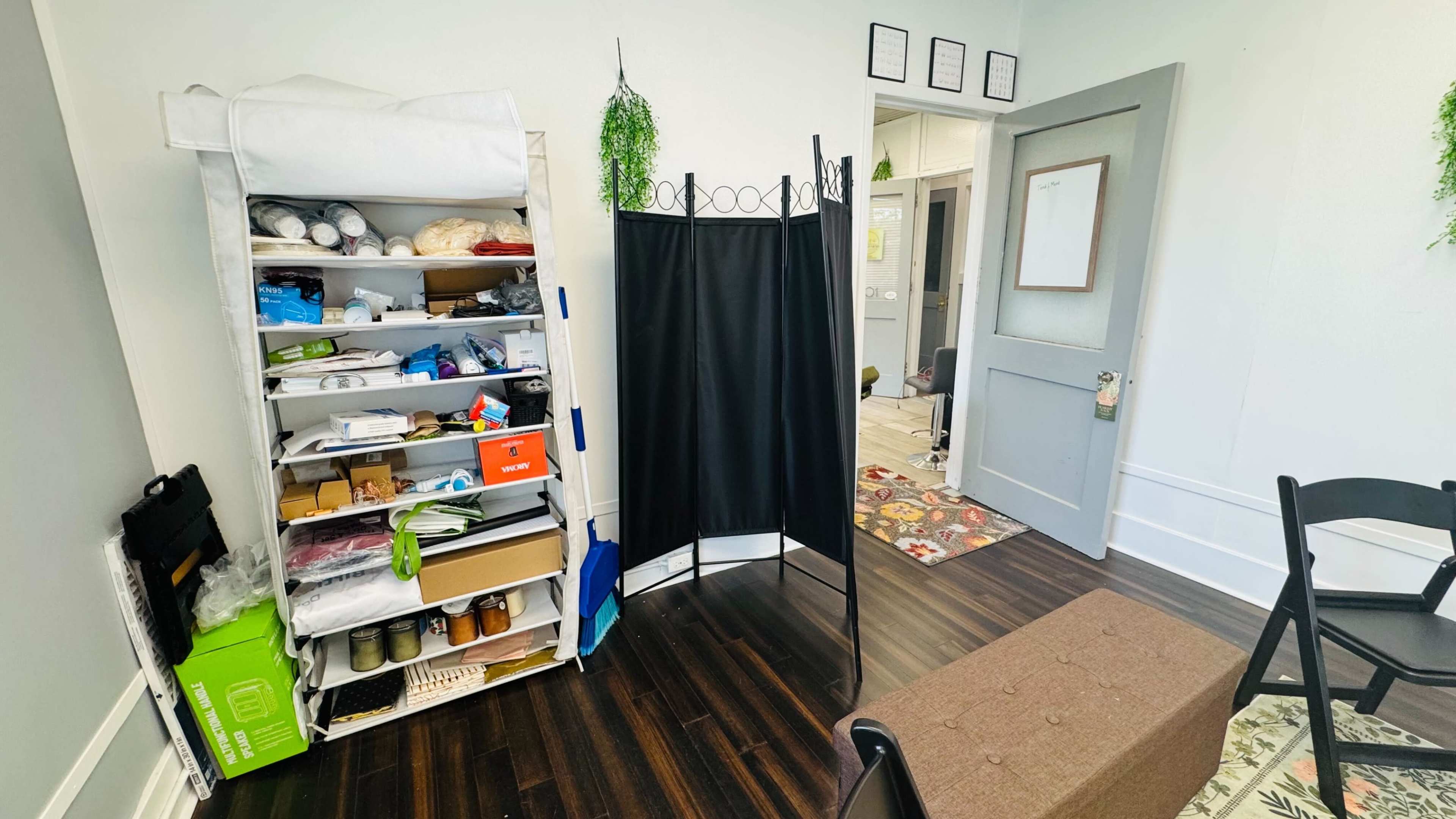 The image shows a room with a shelving unit filled with various supplies, a black folding privacy screen, and a door leading to another area.