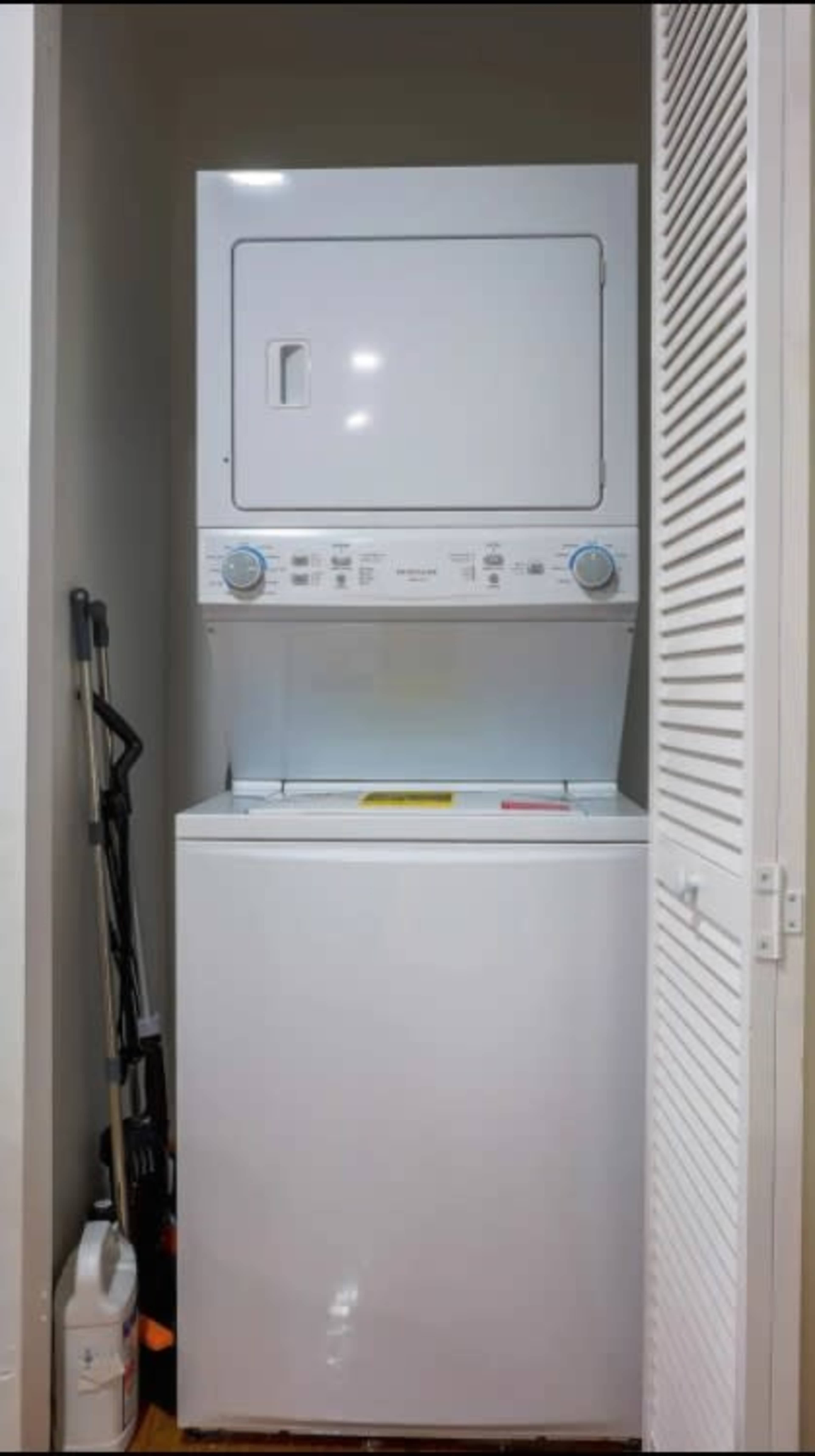 A stacked washer and dryer are located in a closet with a vacuum cleaner and cleaning supplies visible to the side.