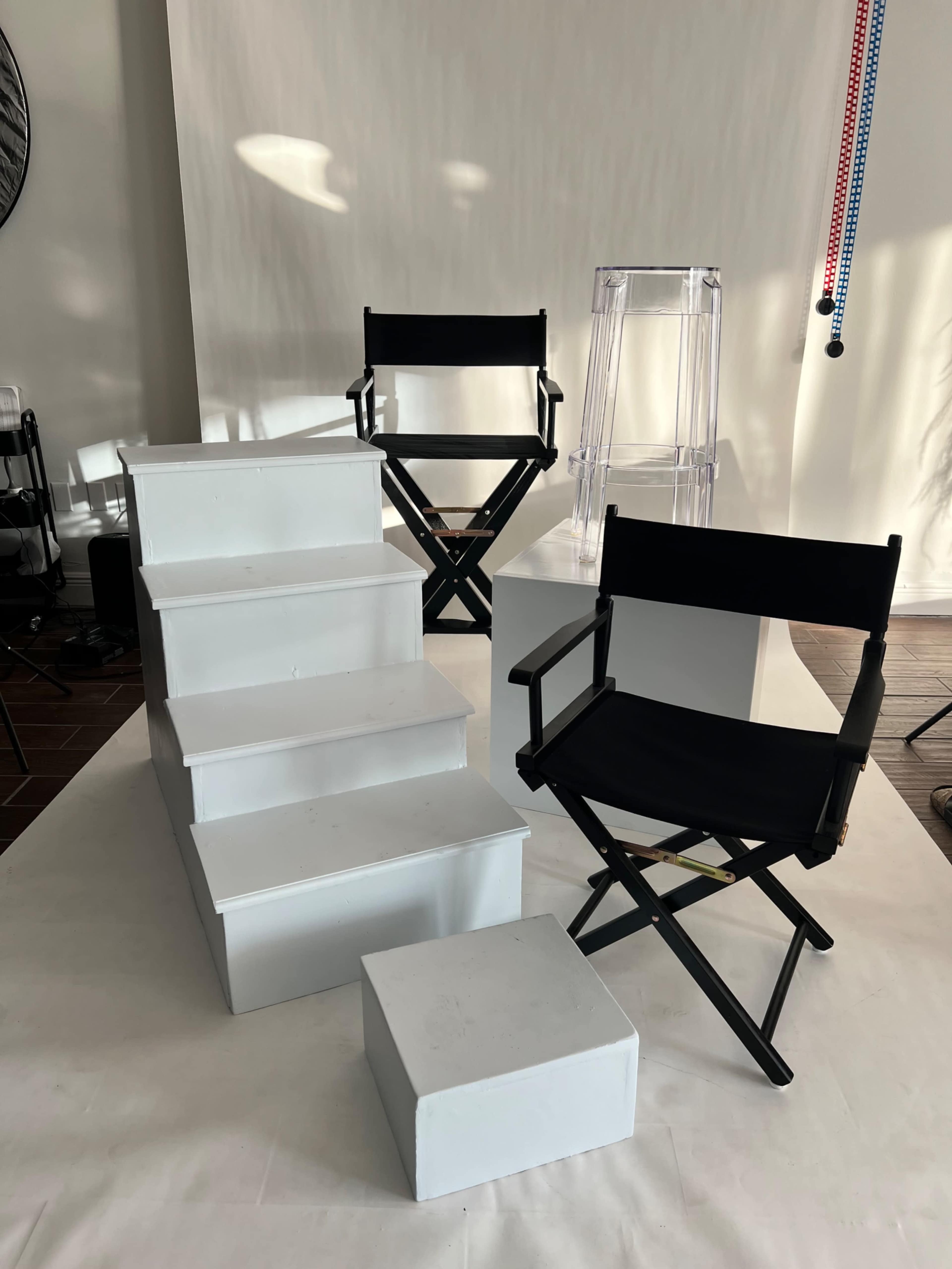The image features a set of white steps with two black director's chairs and a clear stool arranged in a studio setting.