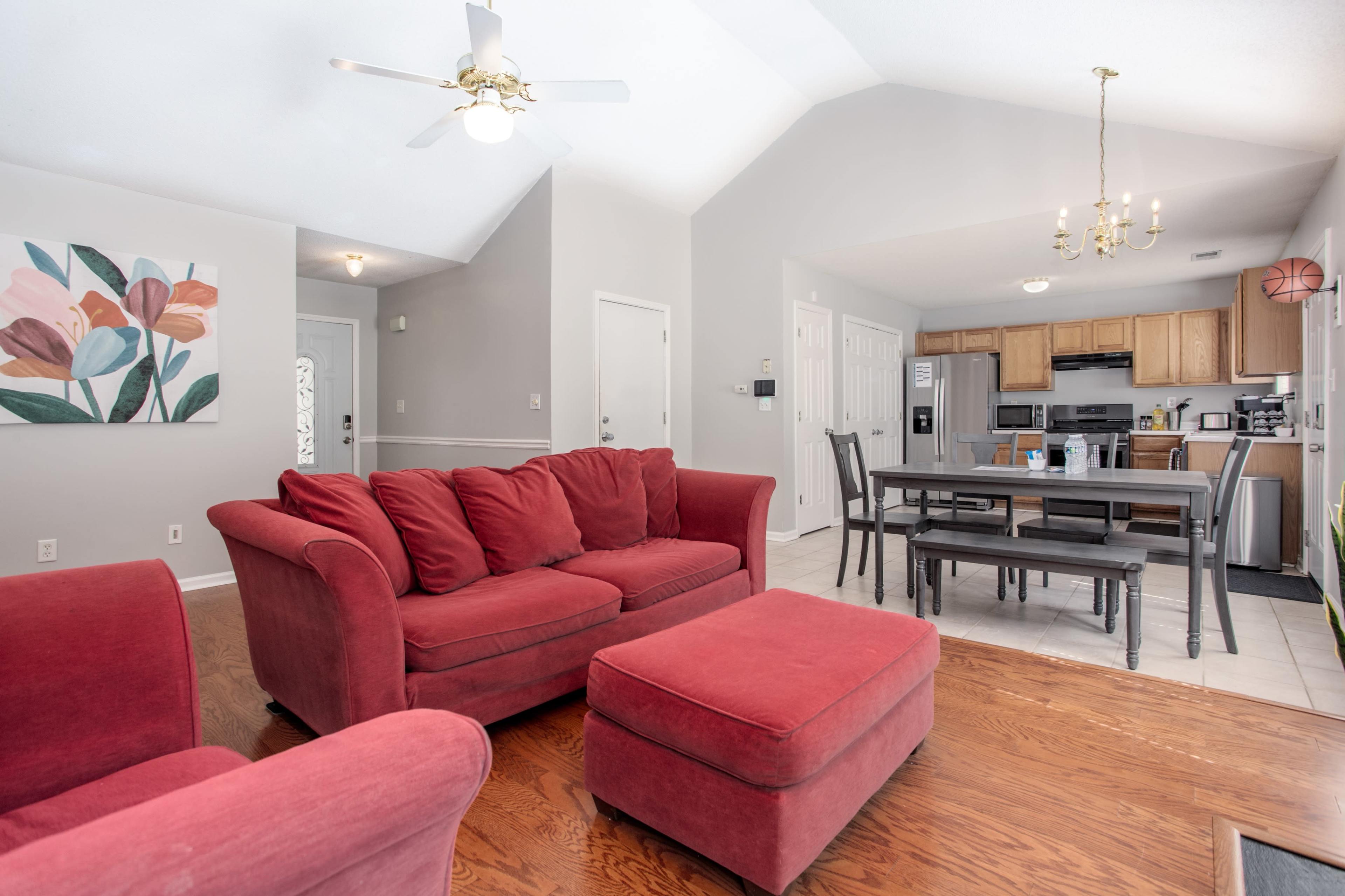 A living area features a red couch and ottoman, a dining table, and an open kitchen with wooden cabinets.