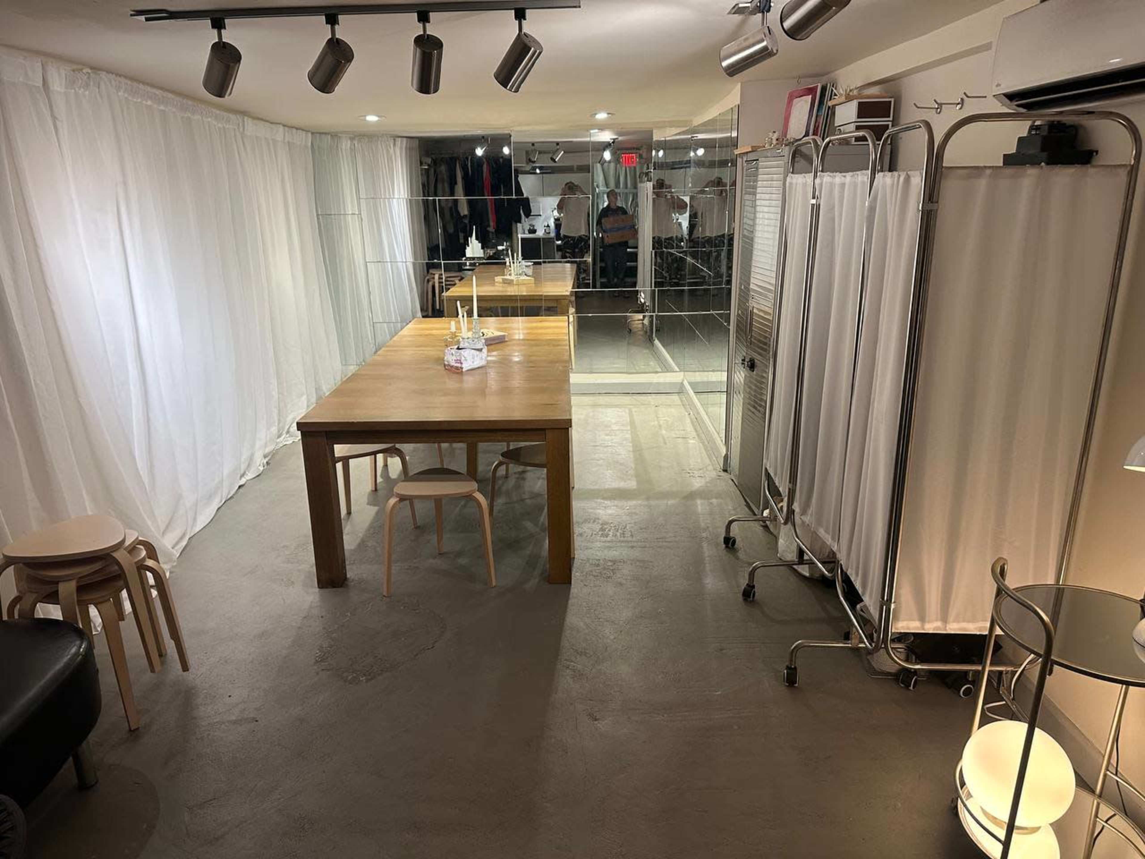 The image shows a spacious room with a wooden table, several chairs, a clothing rack, and partition screens, all set against a backdrop of white drapes.