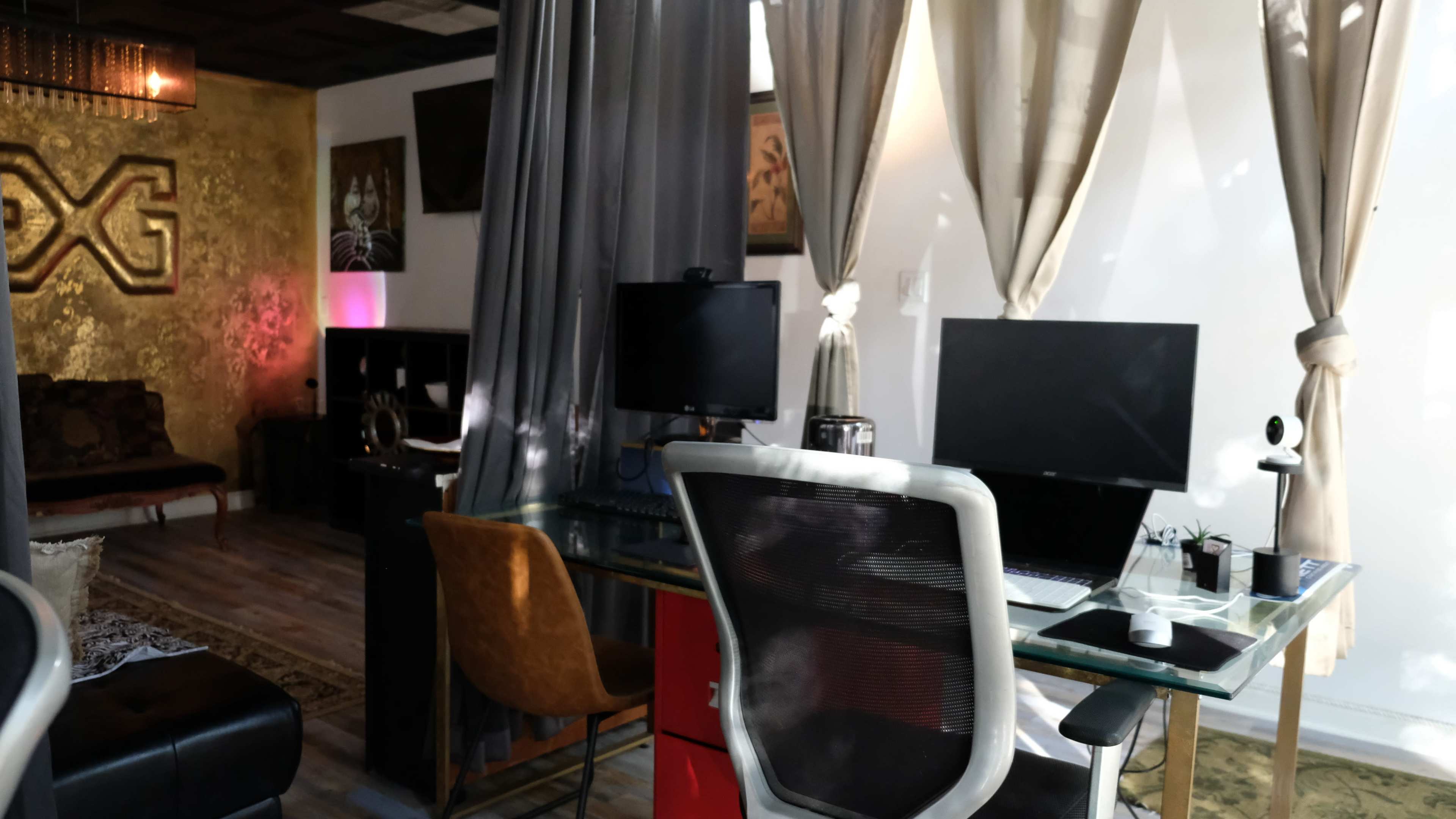 The image shows a modern office space featuring two computer desks, a sofa, and decorative wall art, with sheer curtains and warm lighting.