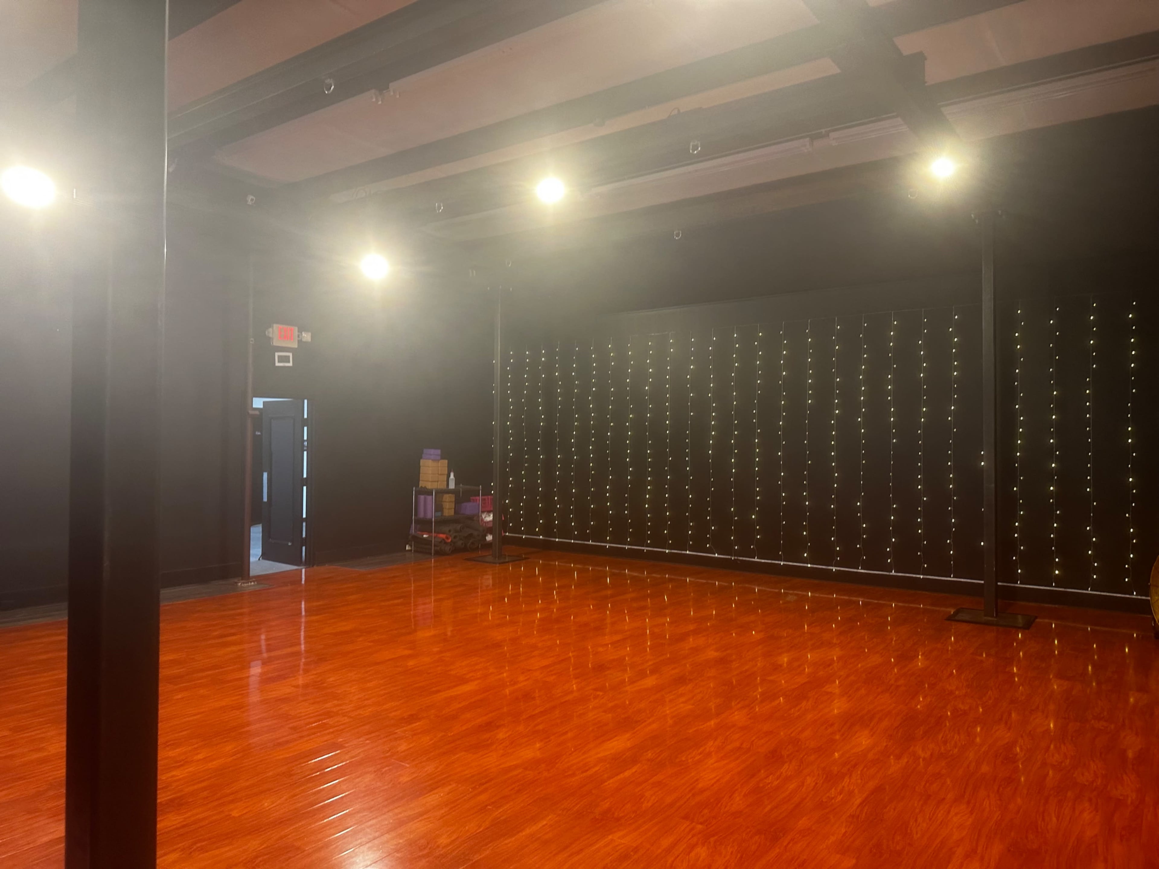 A spacious studio with a shiny wooden floor, illuminated by overhead lights, and a wall adorned with string lights.