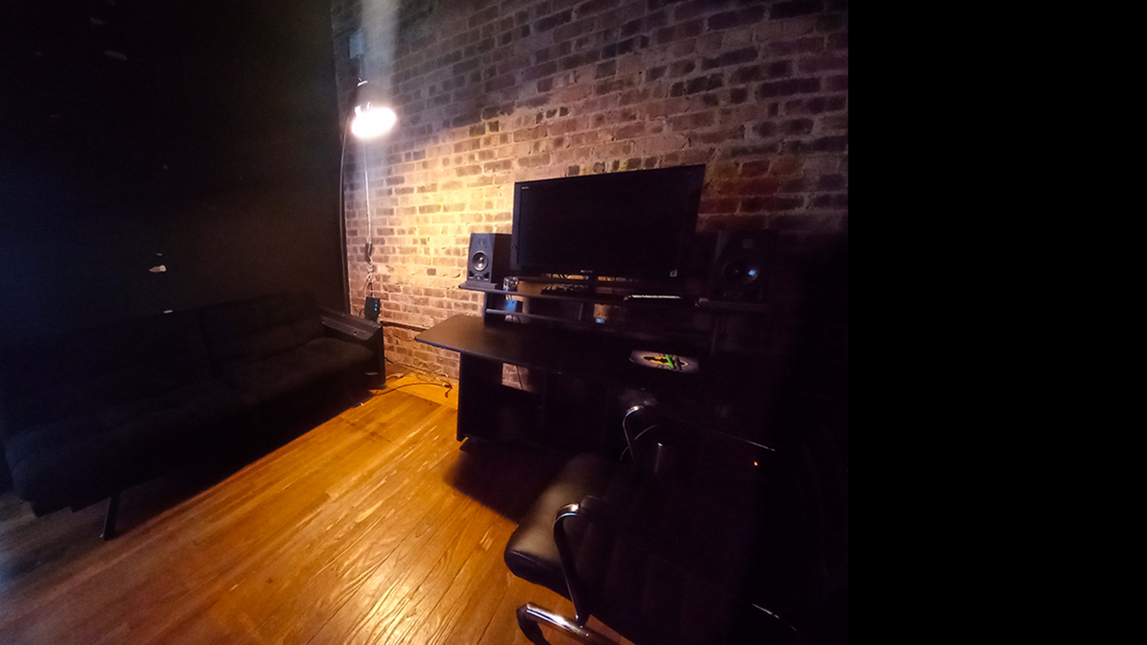 The image shows a dimly lit room with a brick wall, a black sofa, a desk with a computer monitor, and audio speakers.