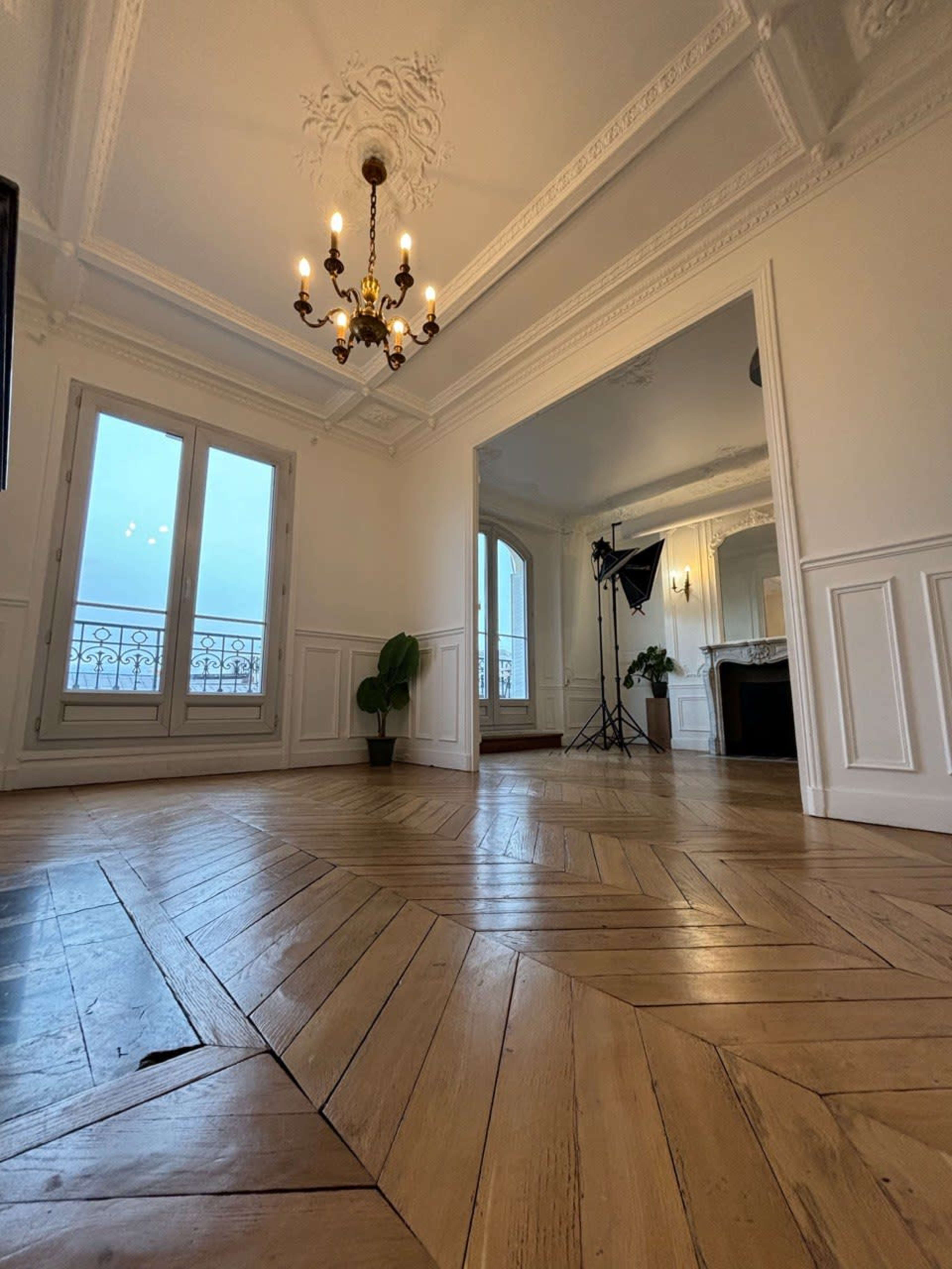 The image shows a well-lit, spacious room with wooden herringbone flooring, elegant molding on the ceiling, and large windows.