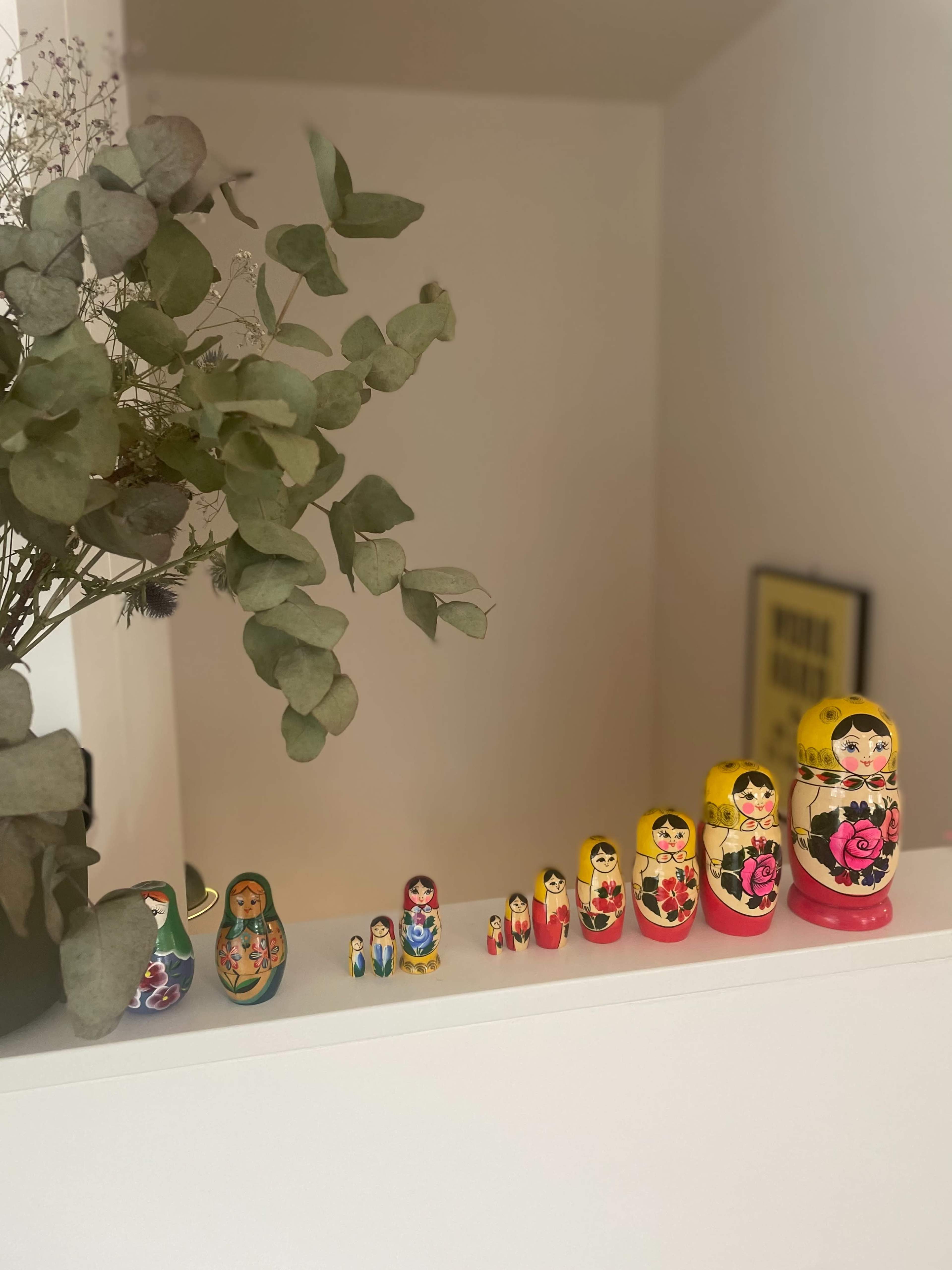 A row of colorful nested Russian dolls, or Matryoshka dolls, is displayed on a white shelf alongside a vase of dried eucalyptus leaves.