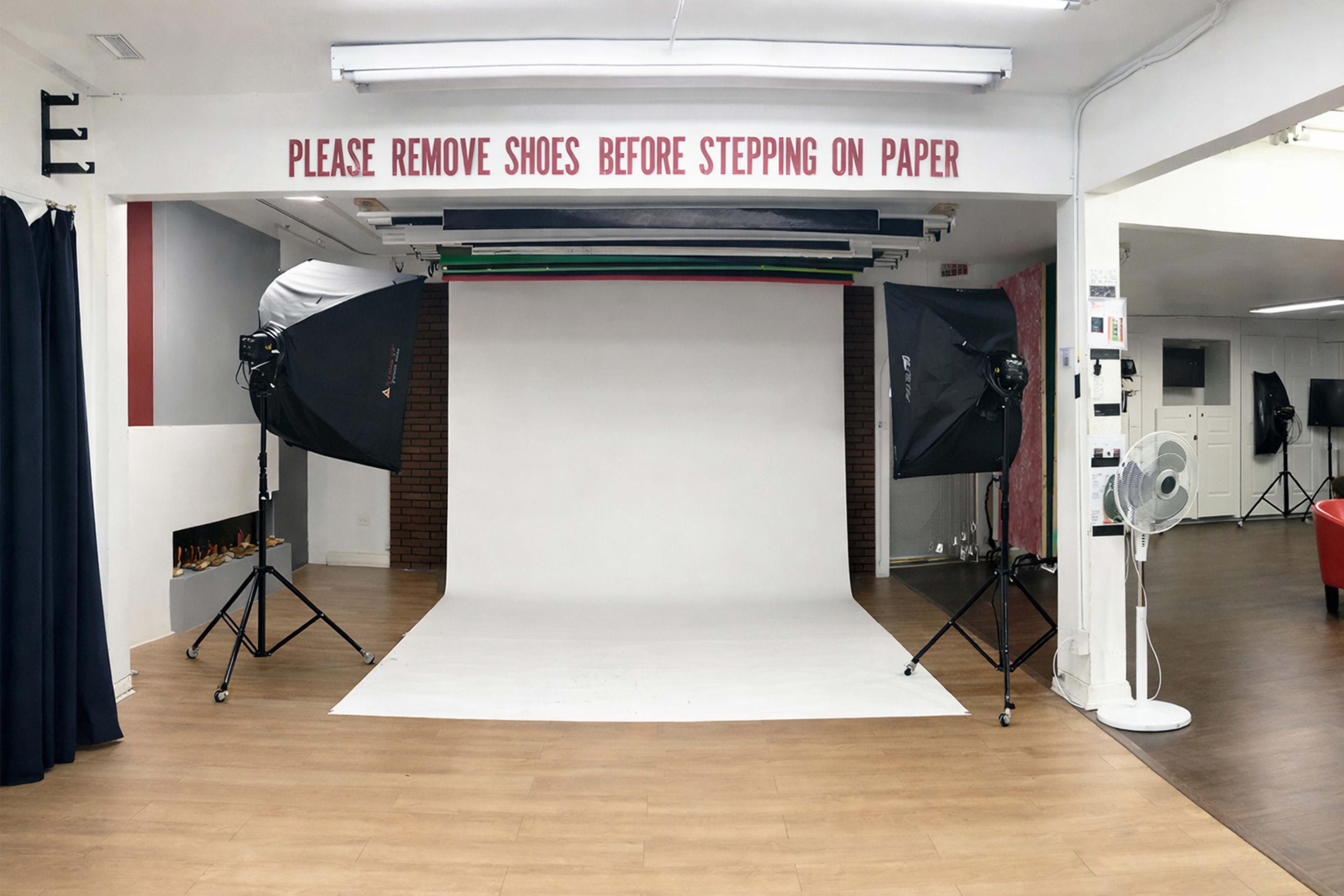 The image shows a photography studio with a white backdrop, softbox lights on either side, and a sign urging to remove shoes before stepping on the paper.