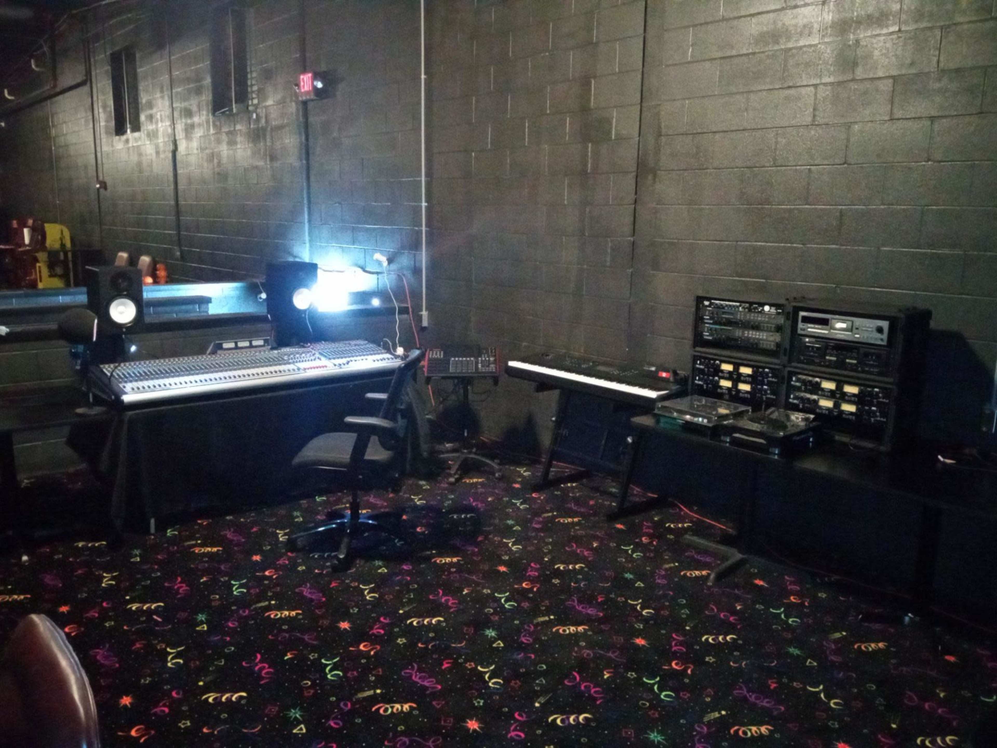 The image shows a music production area featuring a computer mixing console, a keyboard, and audio equipment against a black wall with patterned carpet.