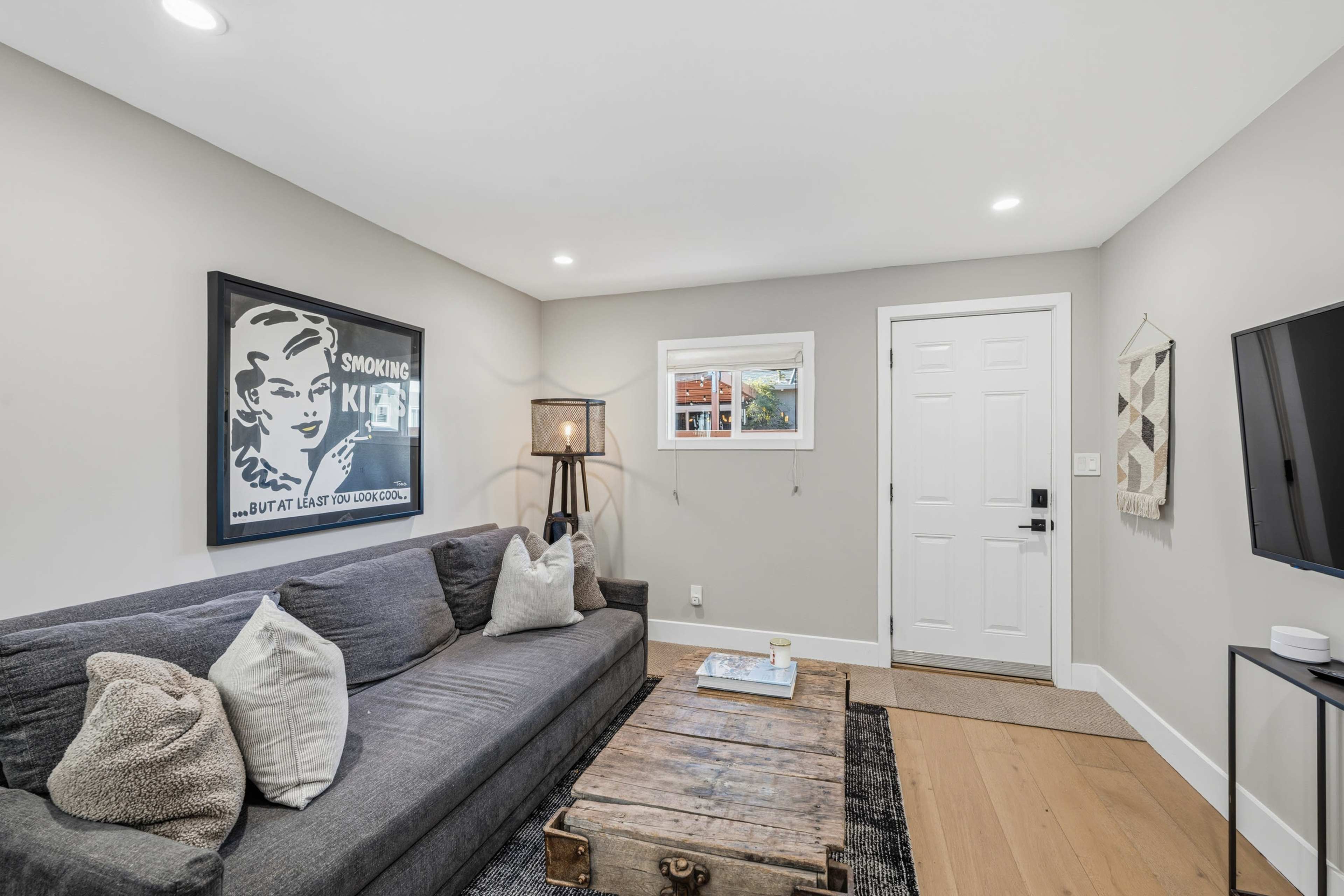 The image shows a modern living room featuring a gray sofa, a wooden coffee table, and a lamp beside the entrance door, with a framed artwork on the wall.