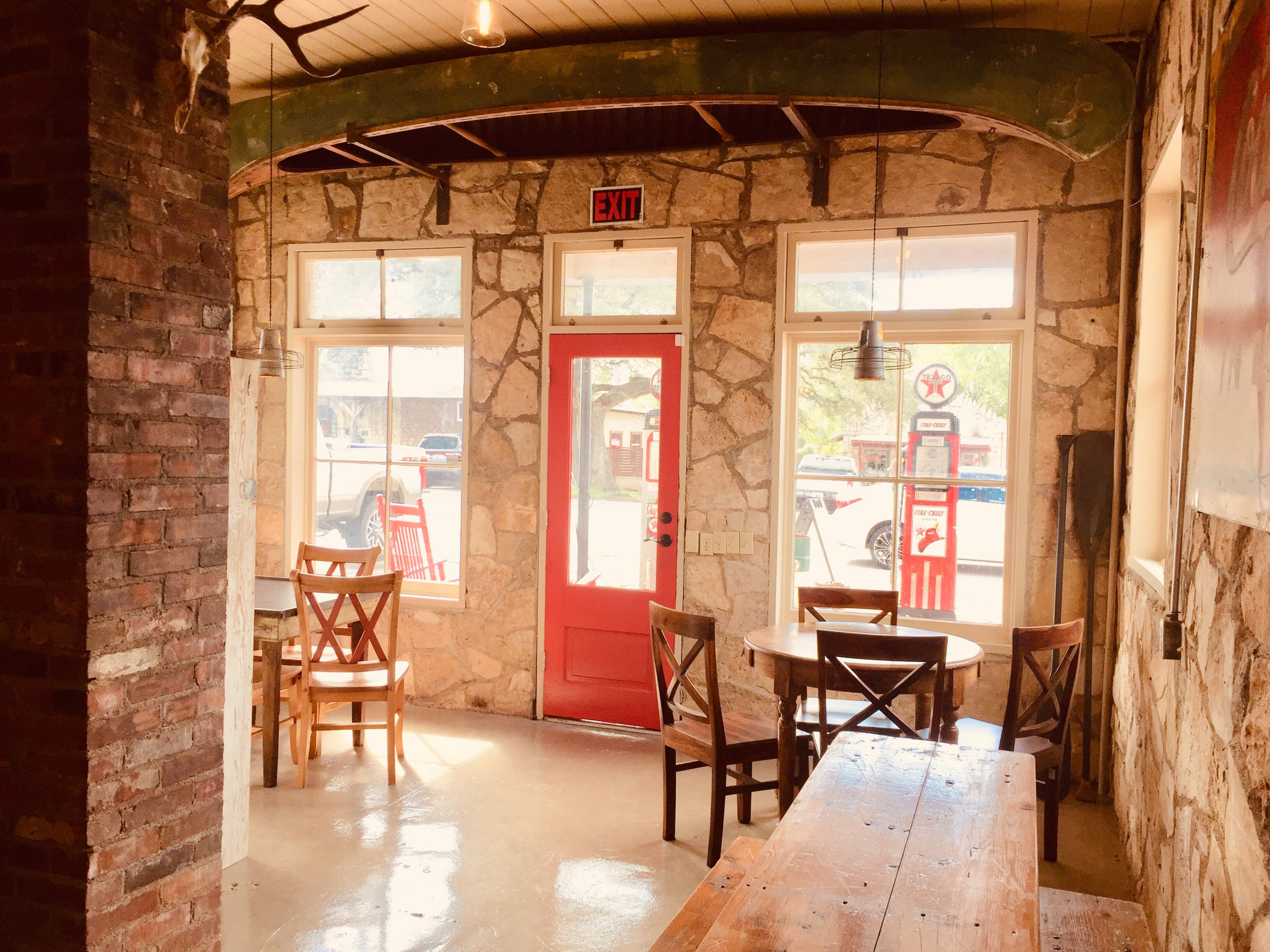 A restaurant interior with stone walls, wooden tables, and chairs, featuring a bright red door and large windows.