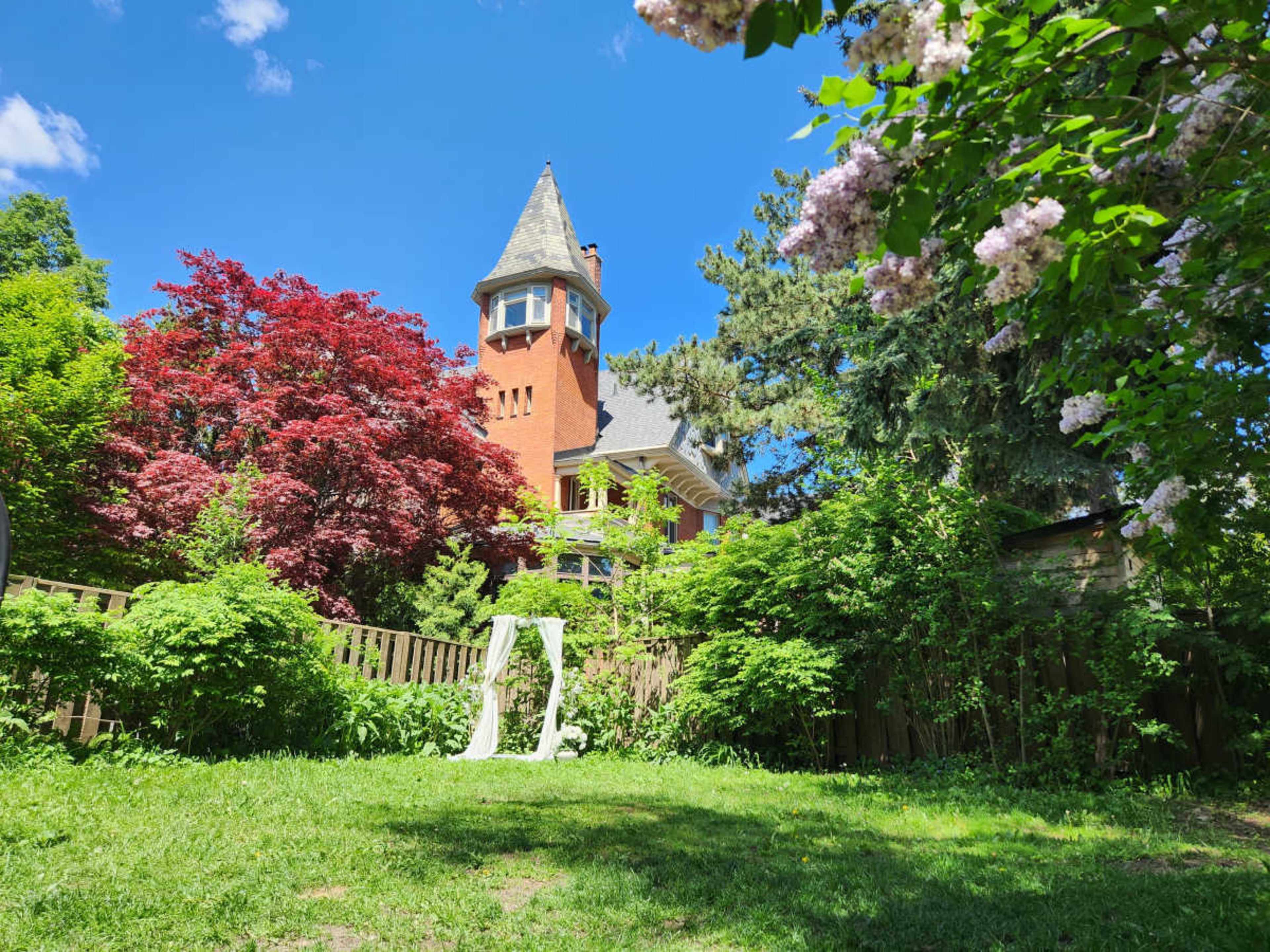 A tall, brick house with a tower is surrounded by lush green foliage and colorful trees in a sunny garden.