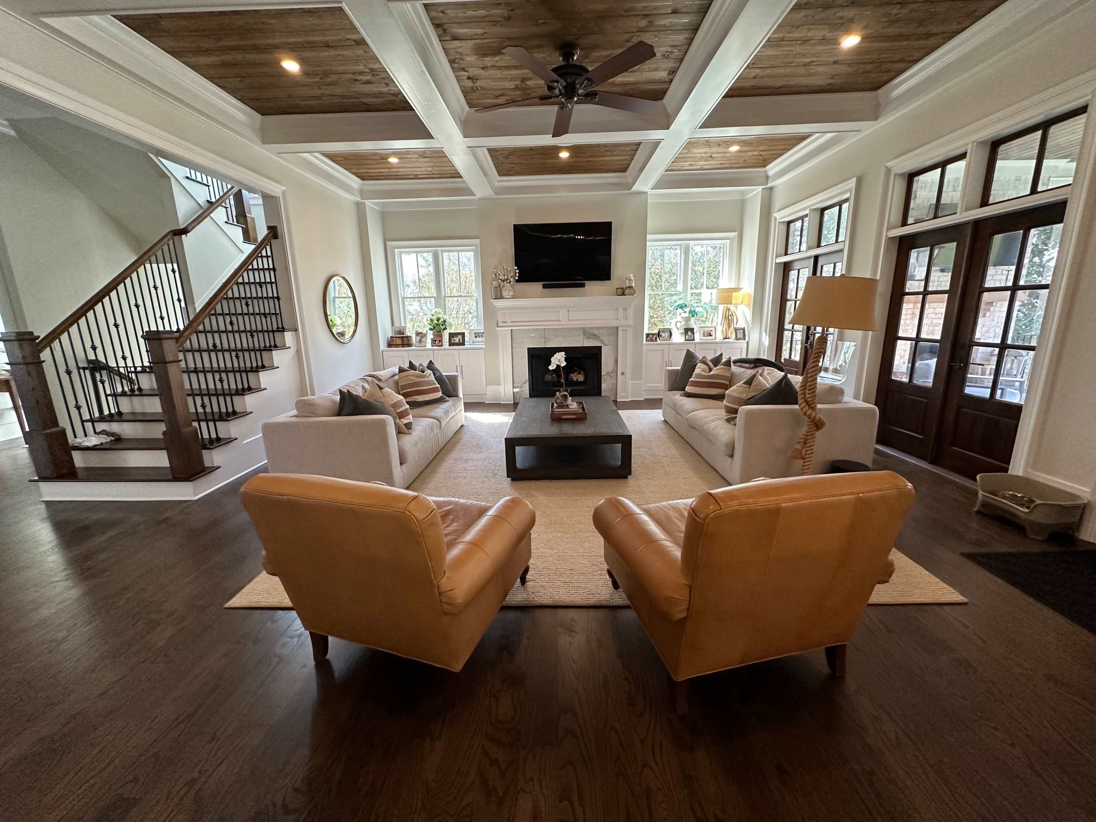 The living room features two cream sofas, two brown leather armchairs, and a central coffee table, with a coffered ceiling and large windows providing natural light.