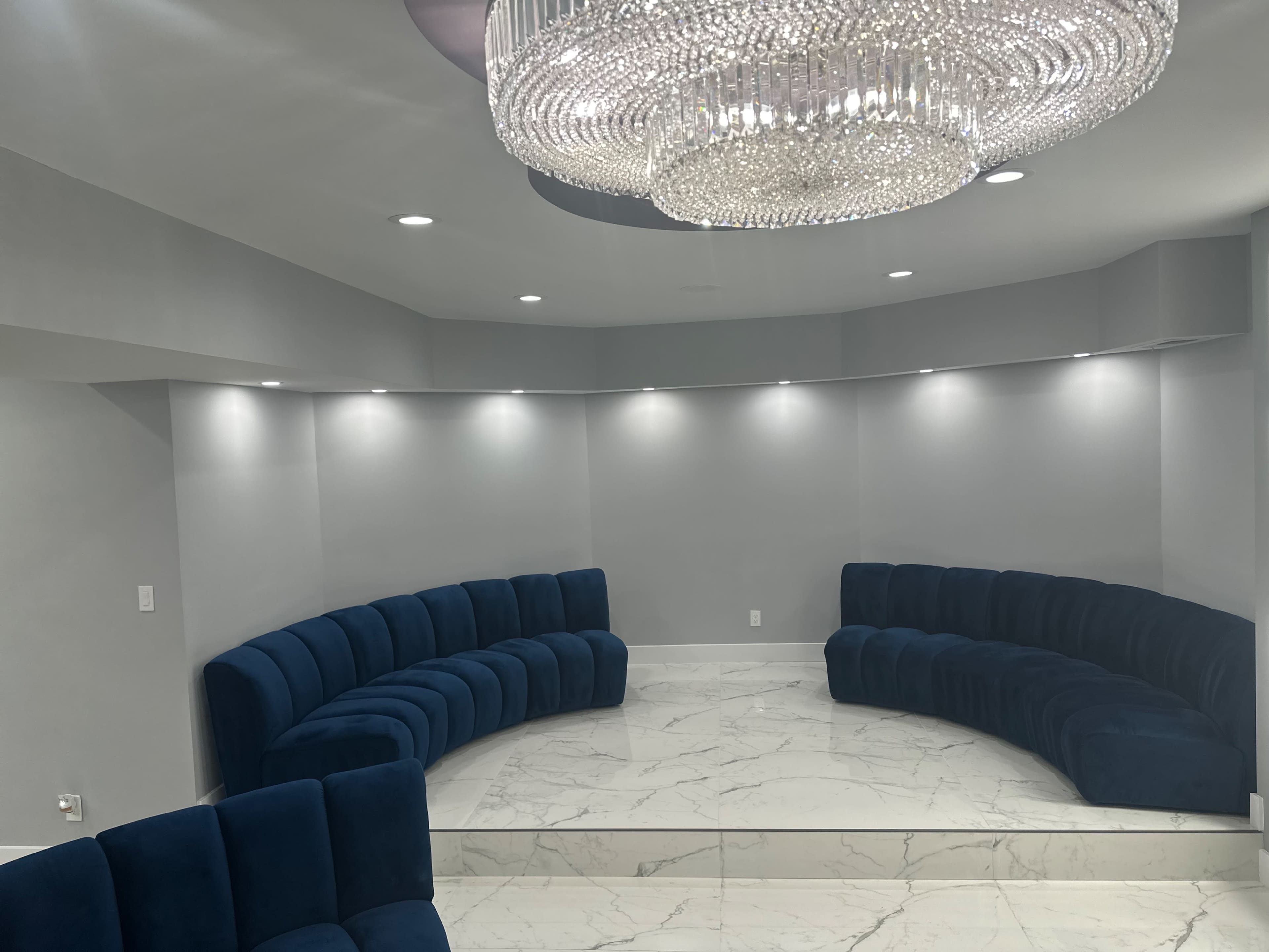 A modern seating area with curved blue sofas arranged around a marble platform, illuminated by a large chandelier overhead.