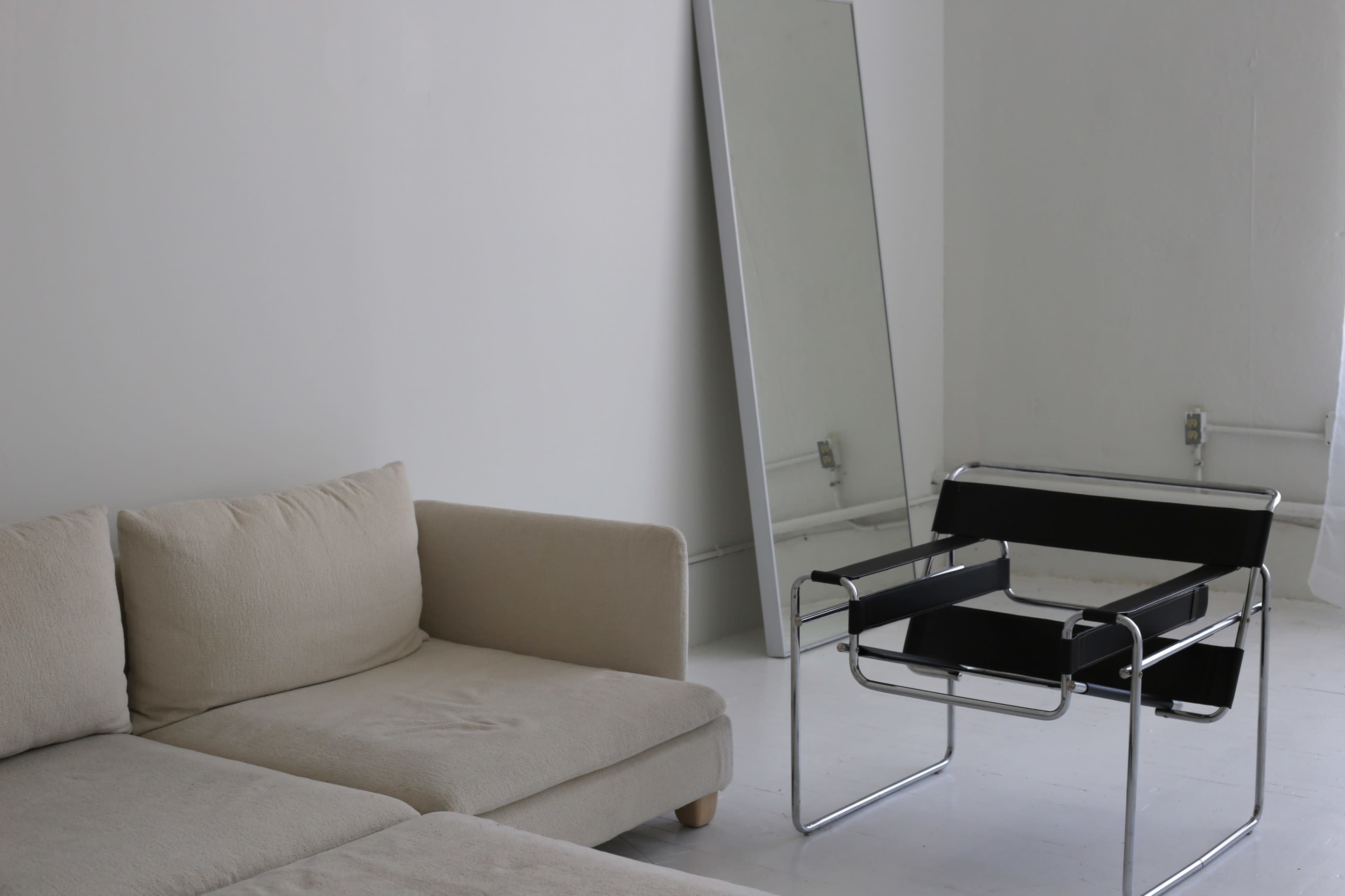 A beige sectional sofa and a black and chrome chair are positioned in a minimally decorated room with a large mirror on the wall.
