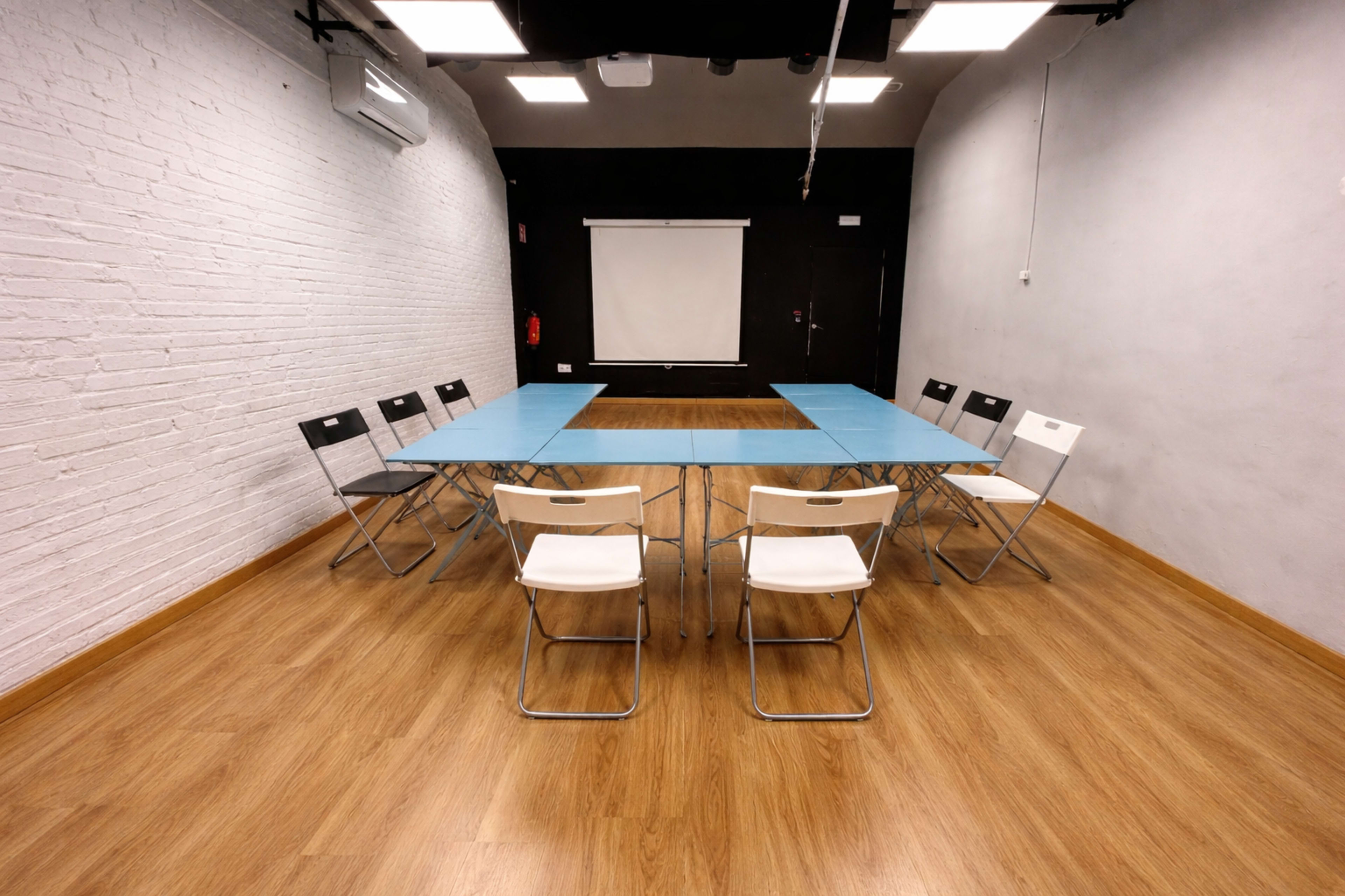 A brightly lit meeting room features a rectangular blue table surrounded by white and black folding chairs, with a blank projection screen on the back wall.