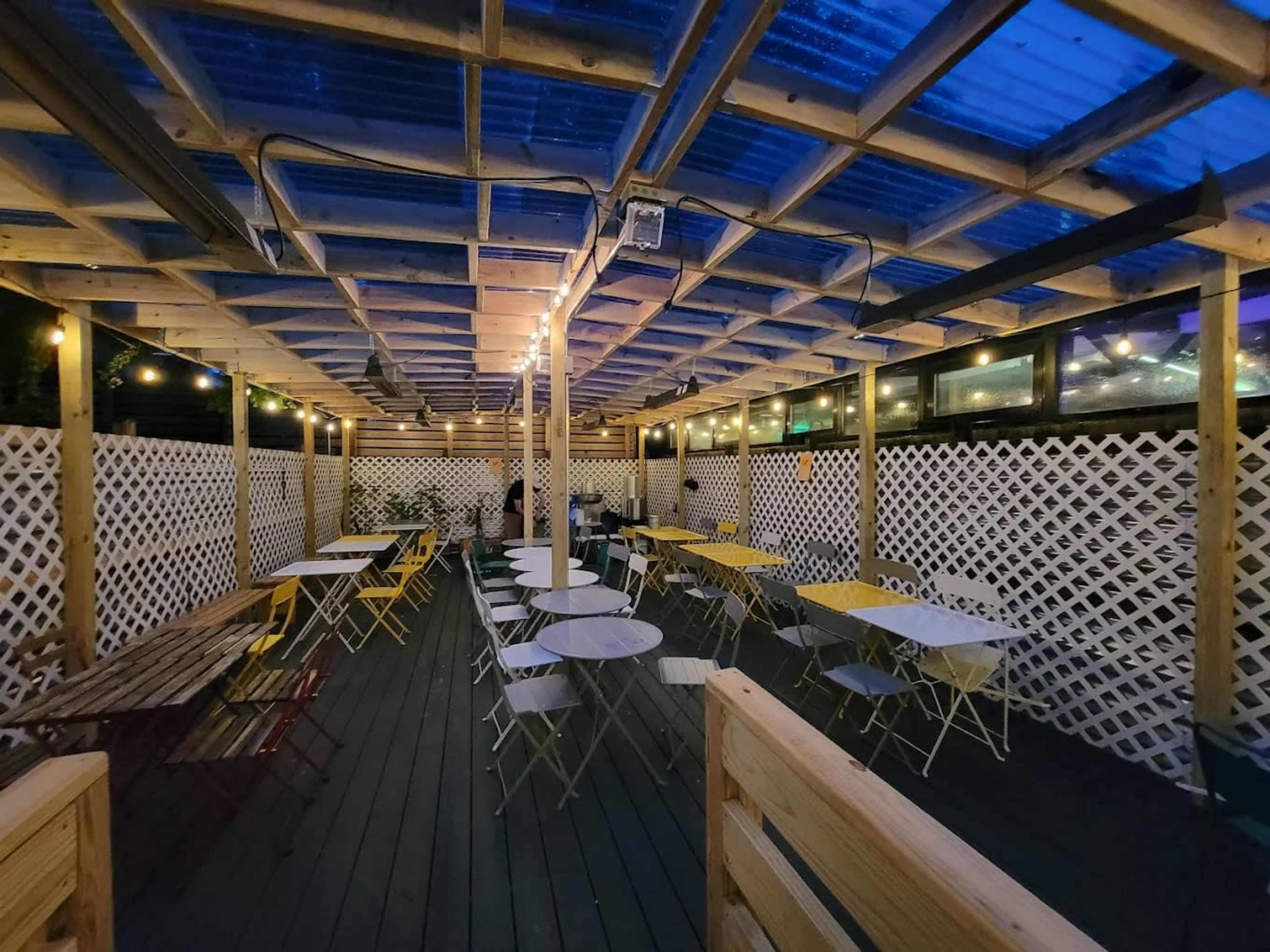 A covered outdoor dining area features wooden tables and chairs arranged under a lattice structure with string lights above.