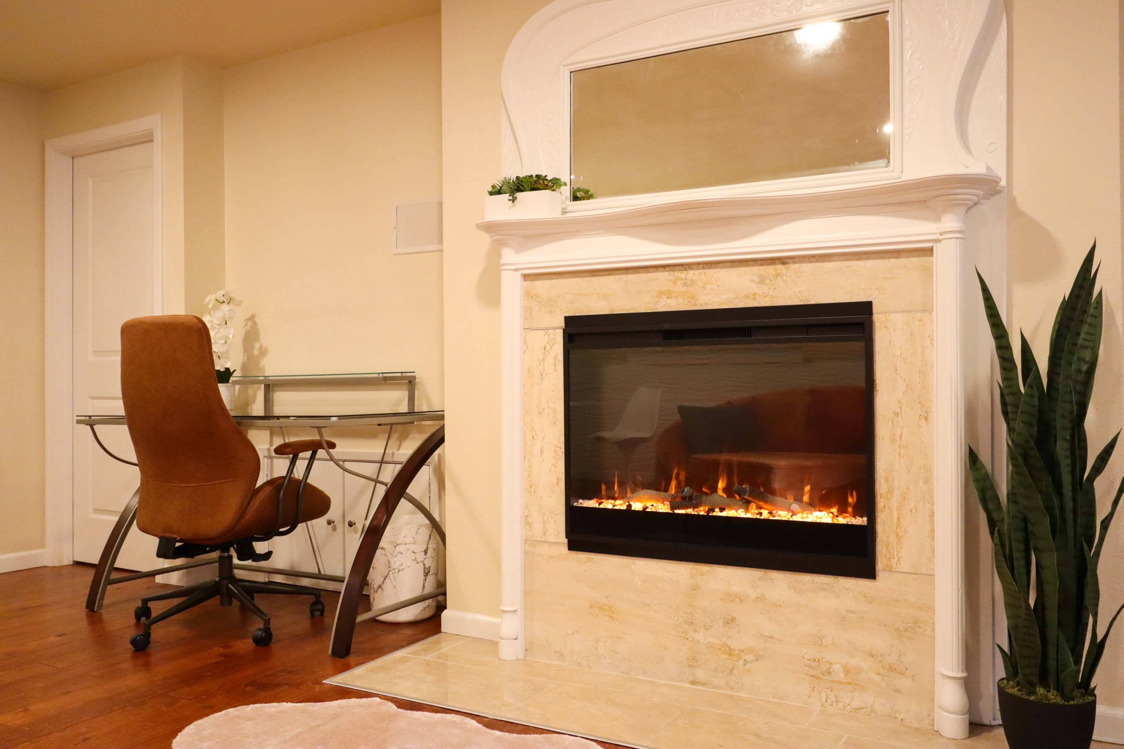 The image shows a cozy interior featuring a modern fireplace set in a neutral-toned wall, accompanied by a glass desk and a brown office chair.