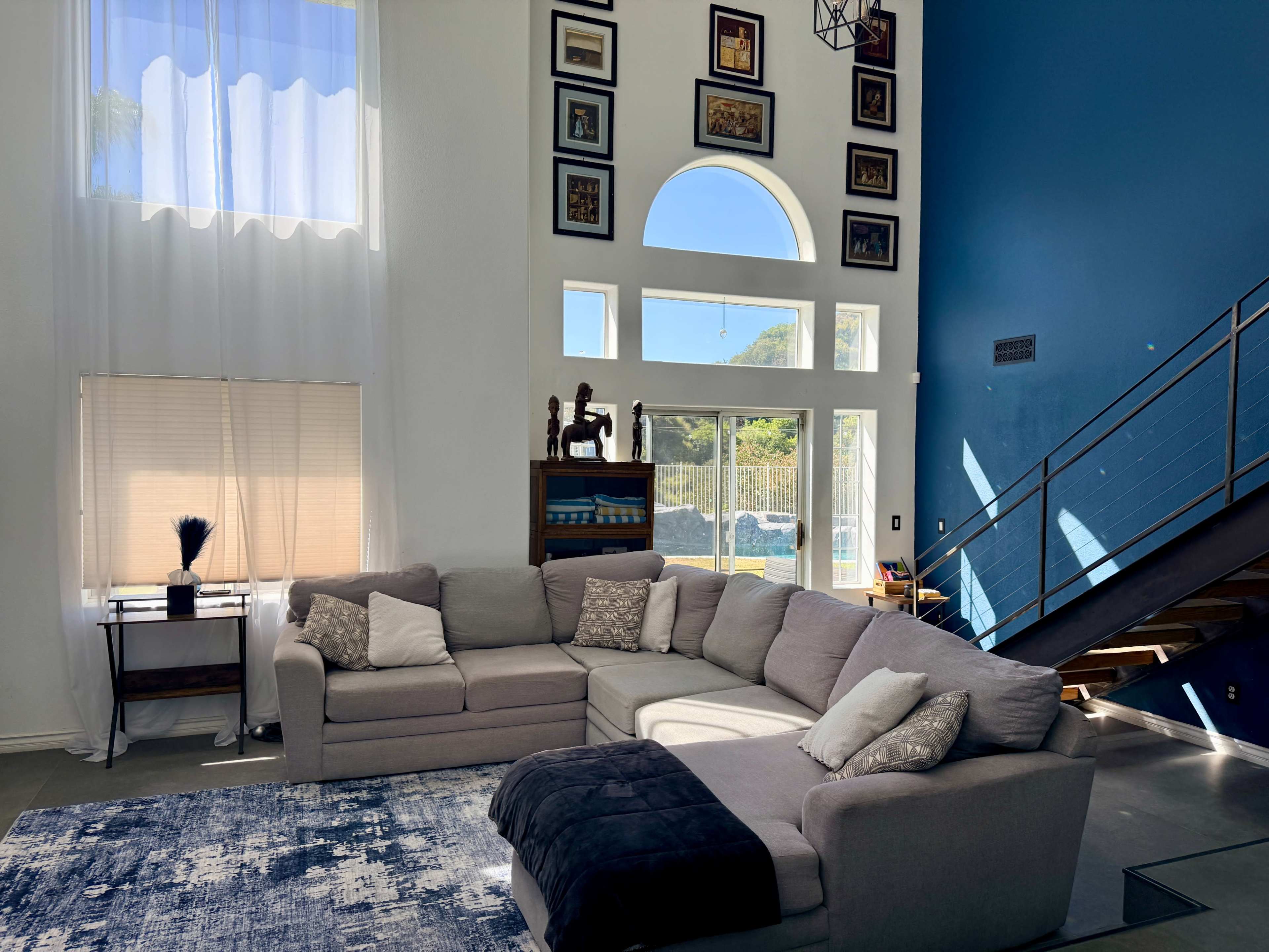 The spacious living room features a large gray sectional sofa, a blue accent wall, and an array of framed artwork displayed above a staircase.