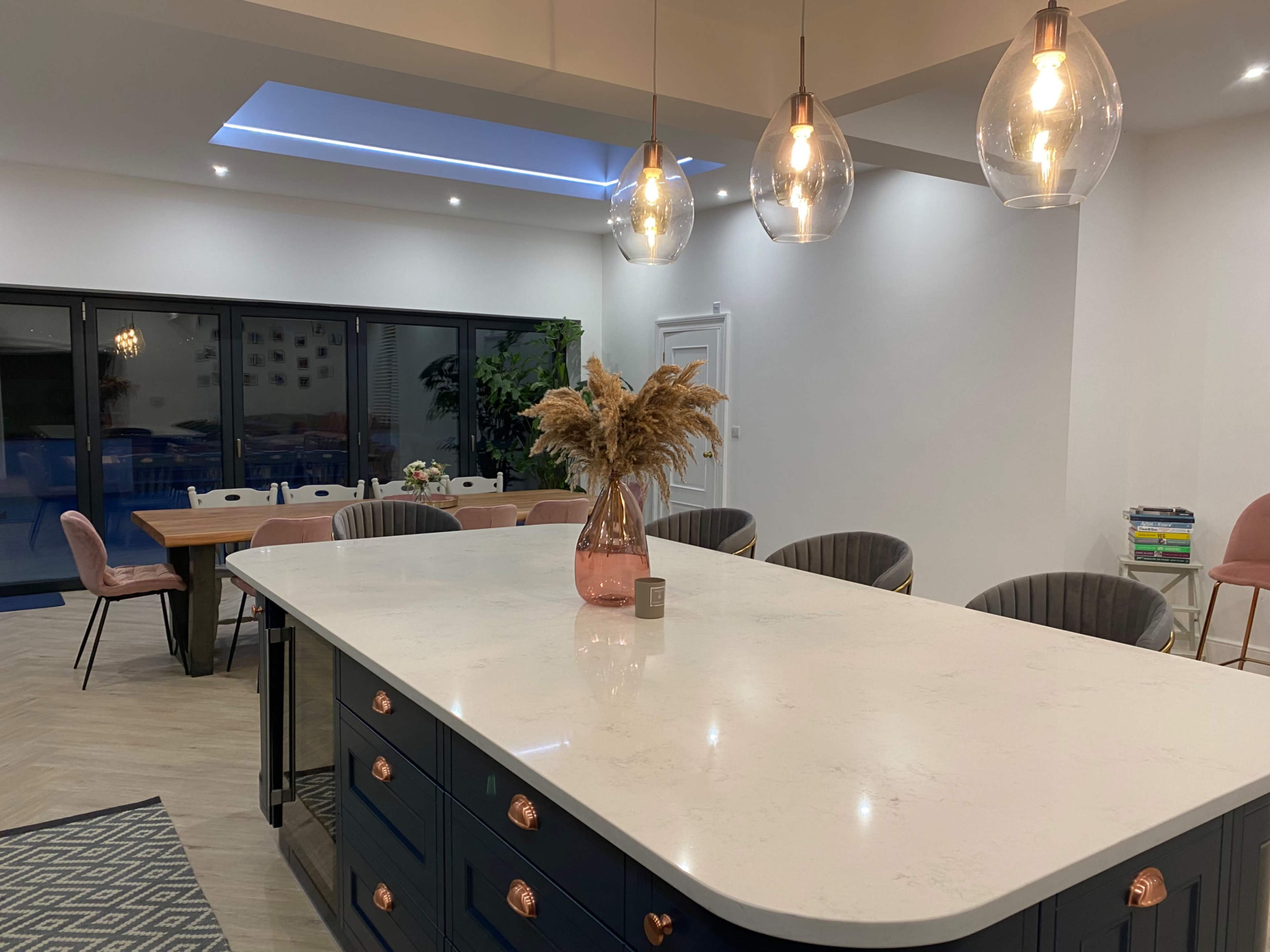 A modern kitchen features a large island with a marble countertop, pendant lights above, and a dining area with wooden chairs in the background.