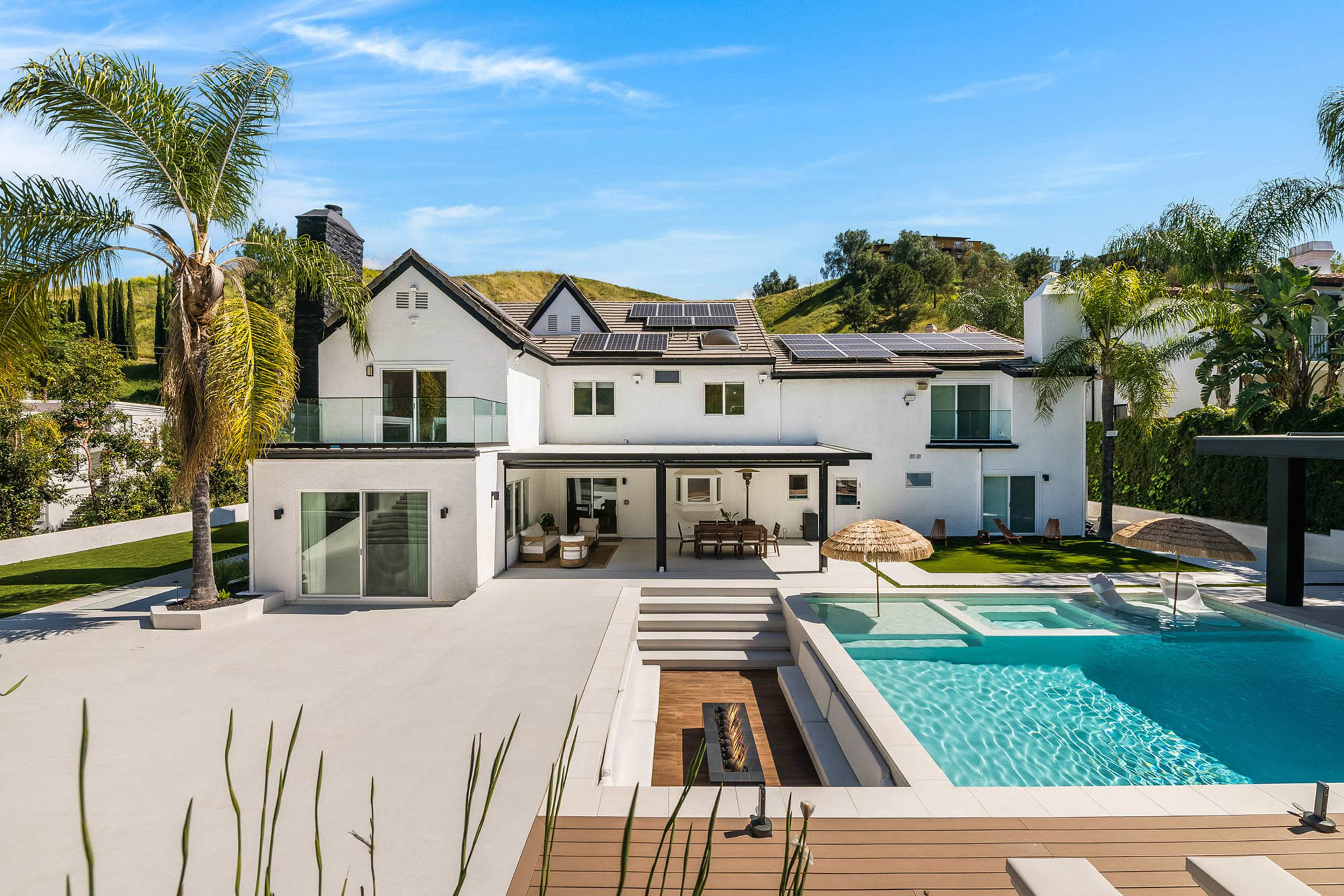 The image shows a modern white house with multiple solar panels on the roof, surrounded by landscaped greenery and a pool with a patio area.