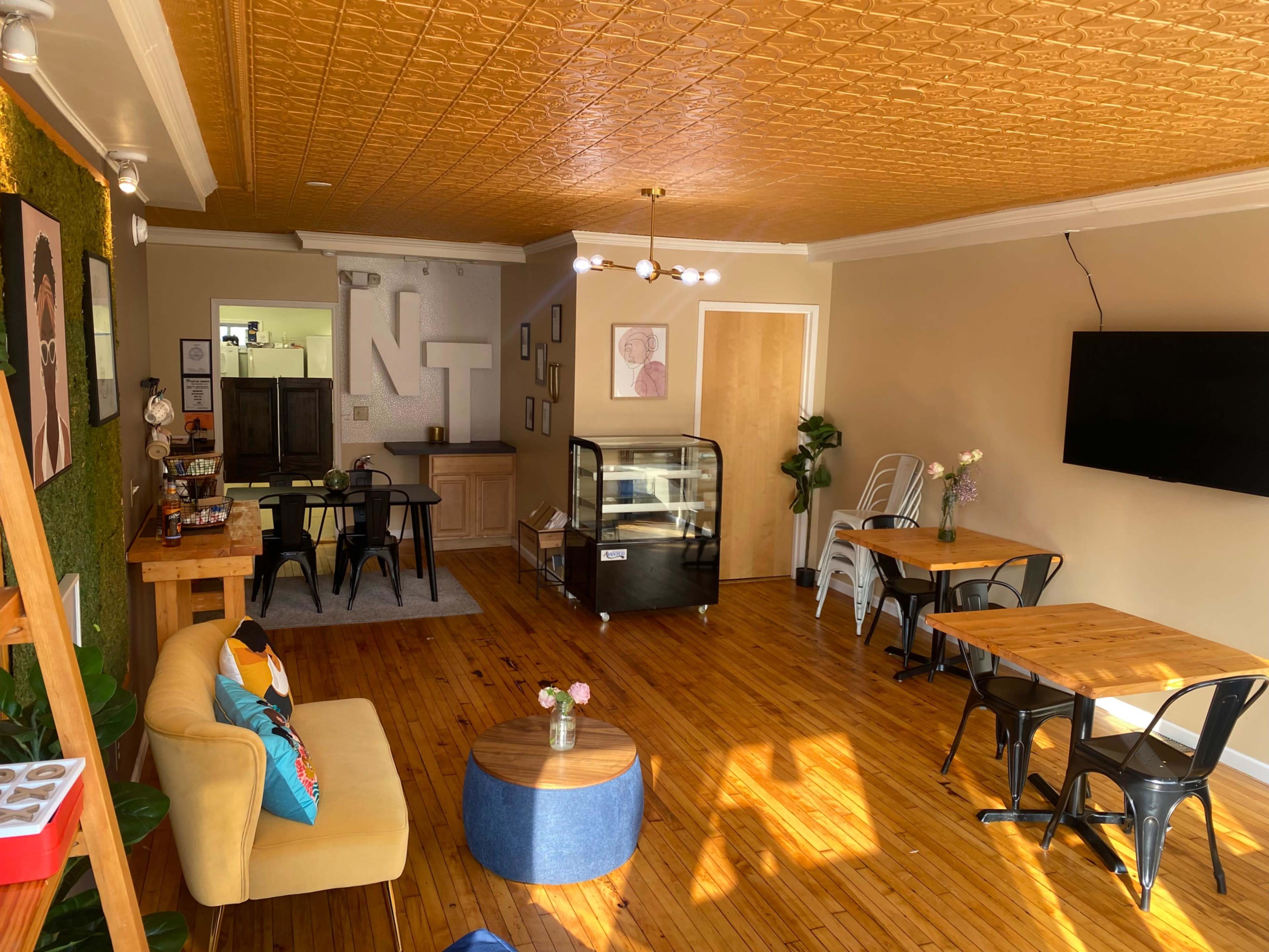 The image shows a cozy café interior with wooden floors, several tables and chairs, a cooler display, and a wall adorned with greenery and artwork.