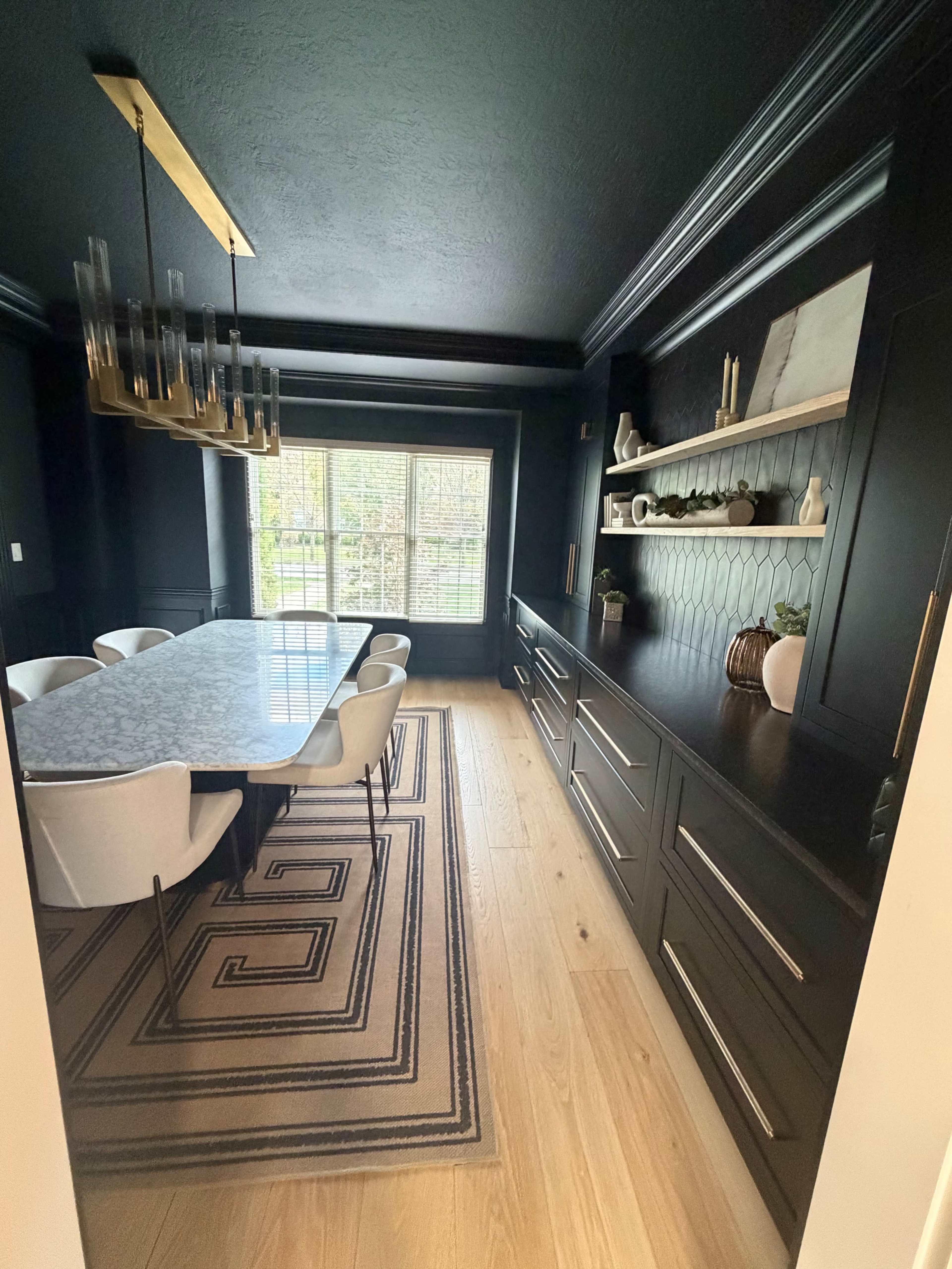 The image depicts a modern dining room with a large rectangular table, sleek white chairs, and a dark color scheme featuring black walls and cabinetry, along with a patterned area rug.