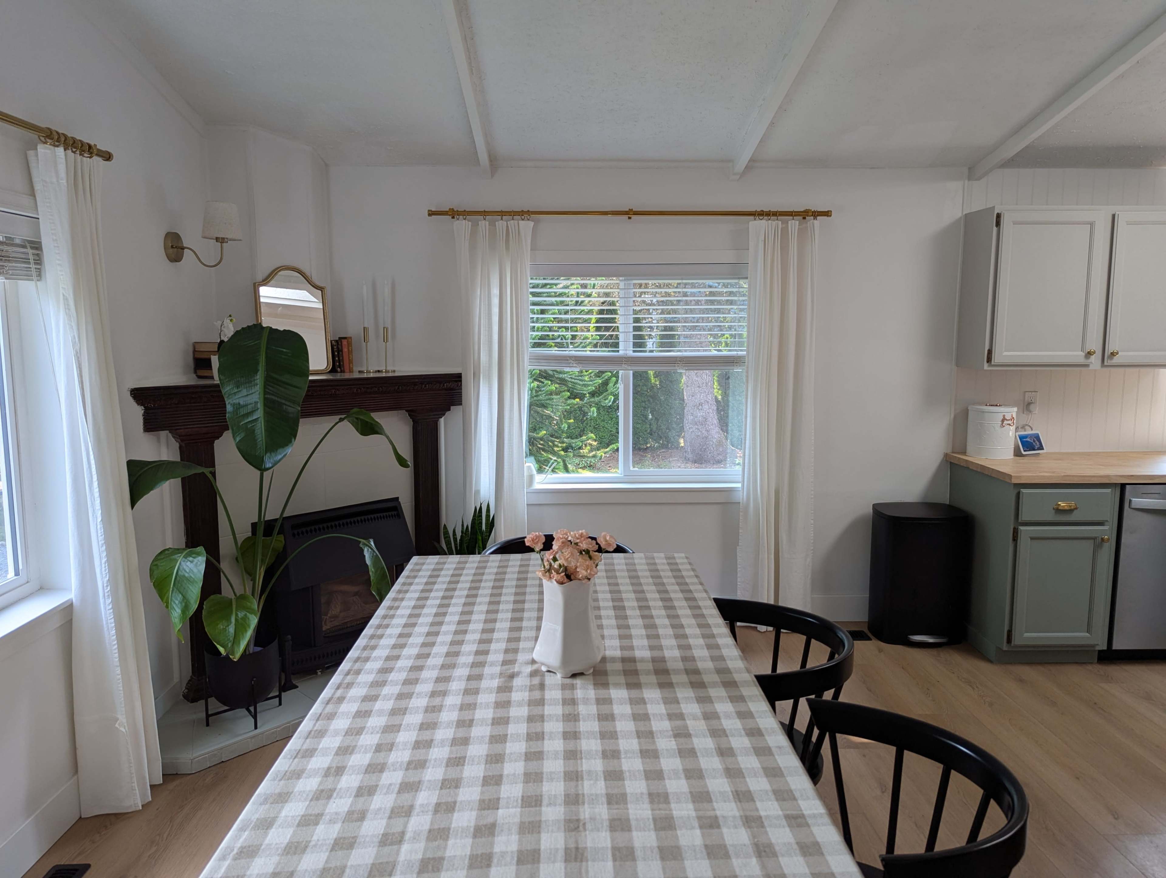 A dining table with a checkered tablecloth is positioned in a bright kitchen, which features a large window and a green cabinet.