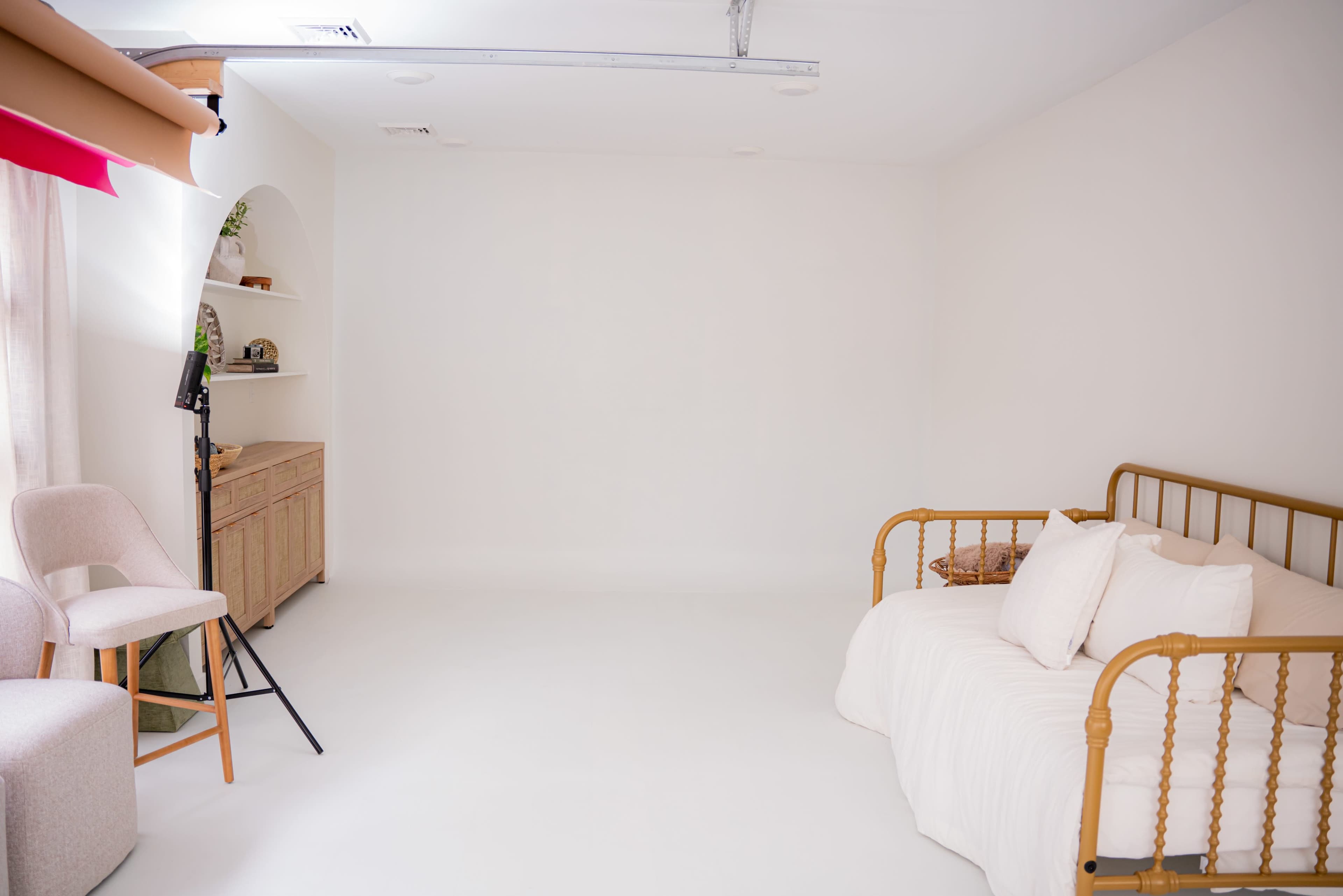 The image shows a minimalist interior space featuring a light-colored sofa bed, a chair, and a shelving unit against a blank wall.