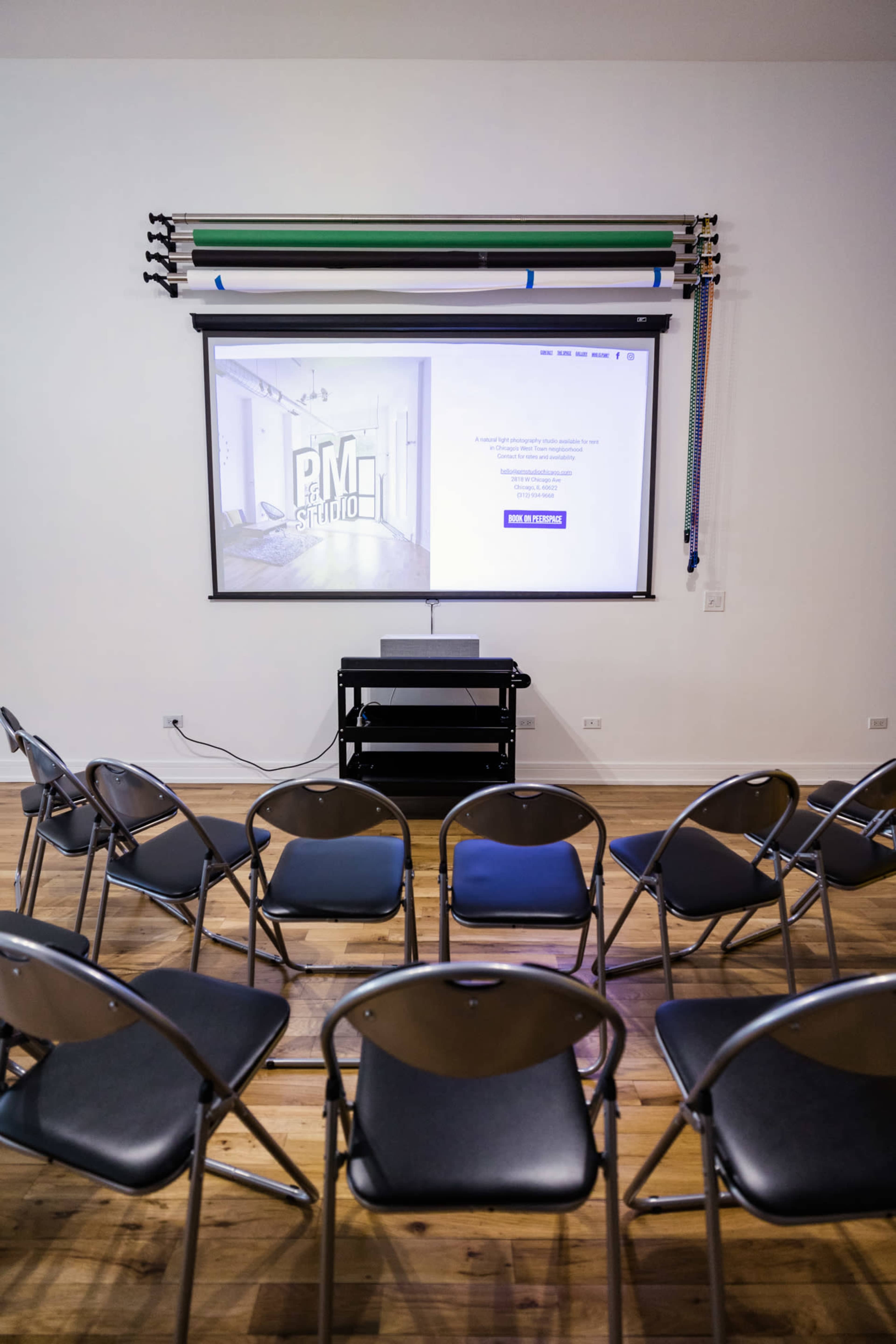 A row of empty folding chairs faces a projection screen displaying a website in a brightly lit room.