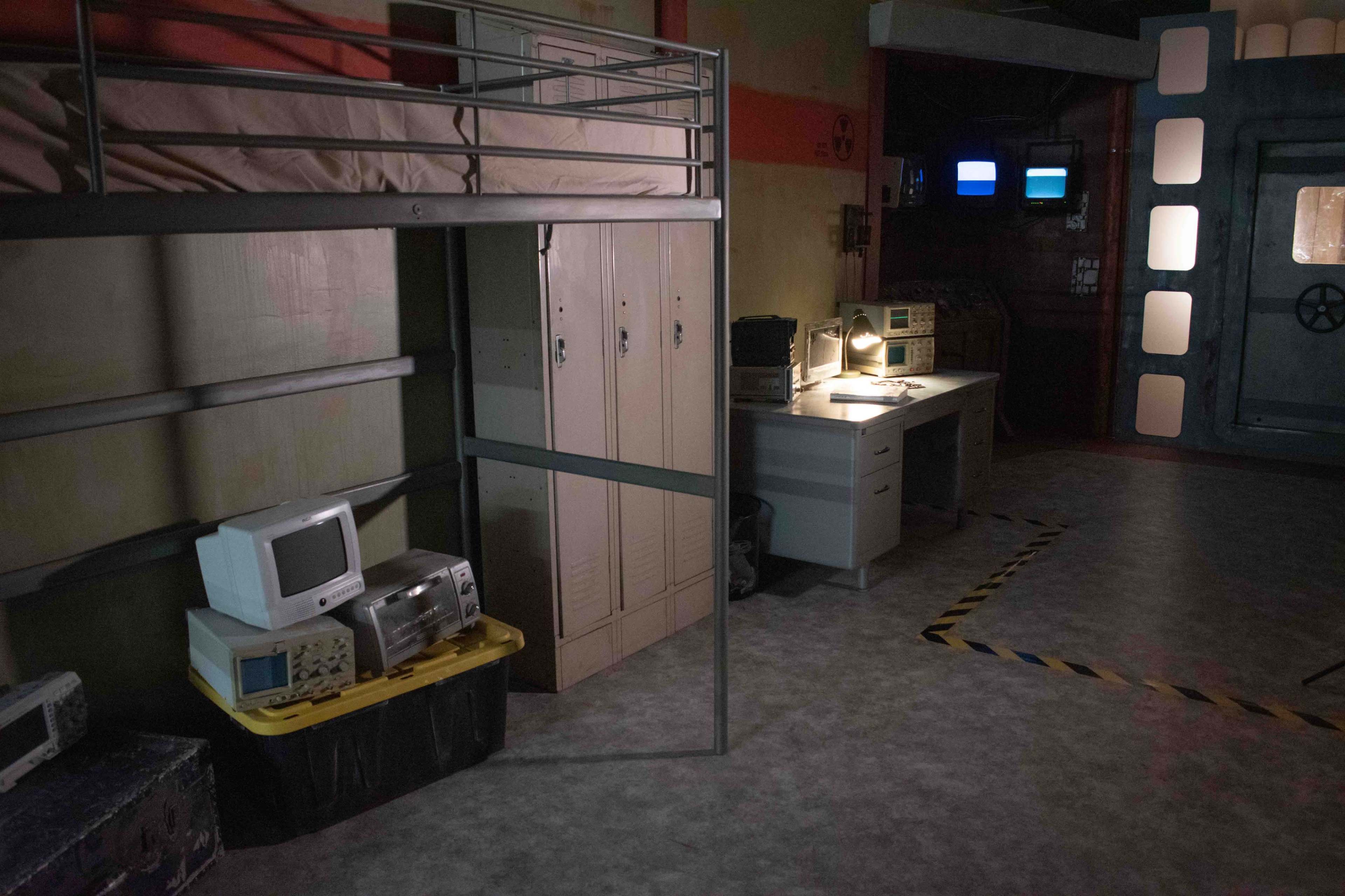 A dimly lit room with a lofted bed, lockers, an old computer, and a desk with a light and monitors.