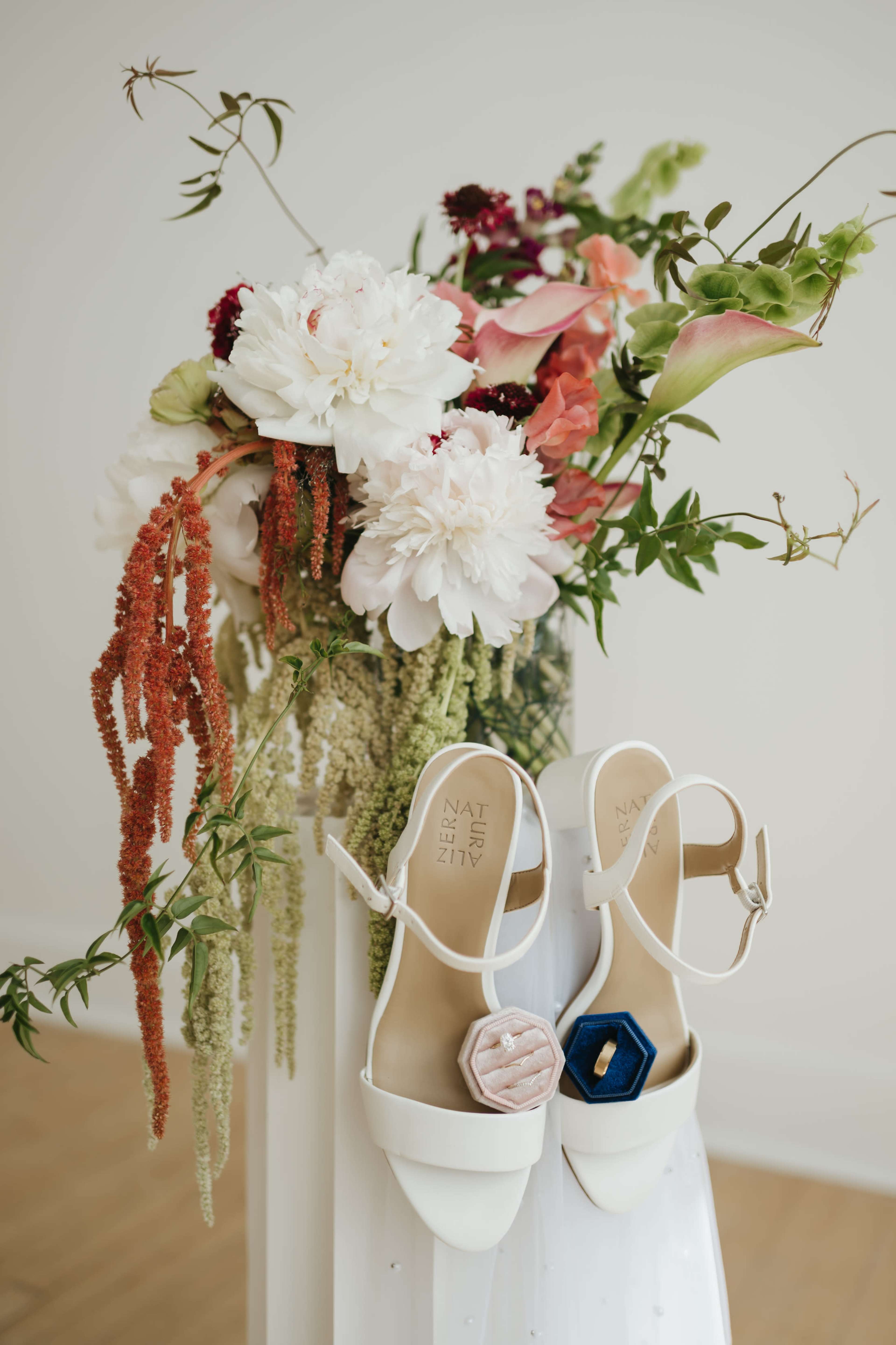 A pair of white sandals with decorative accents is placed beside a tall floral arrangement featuring peonies and other blooms.