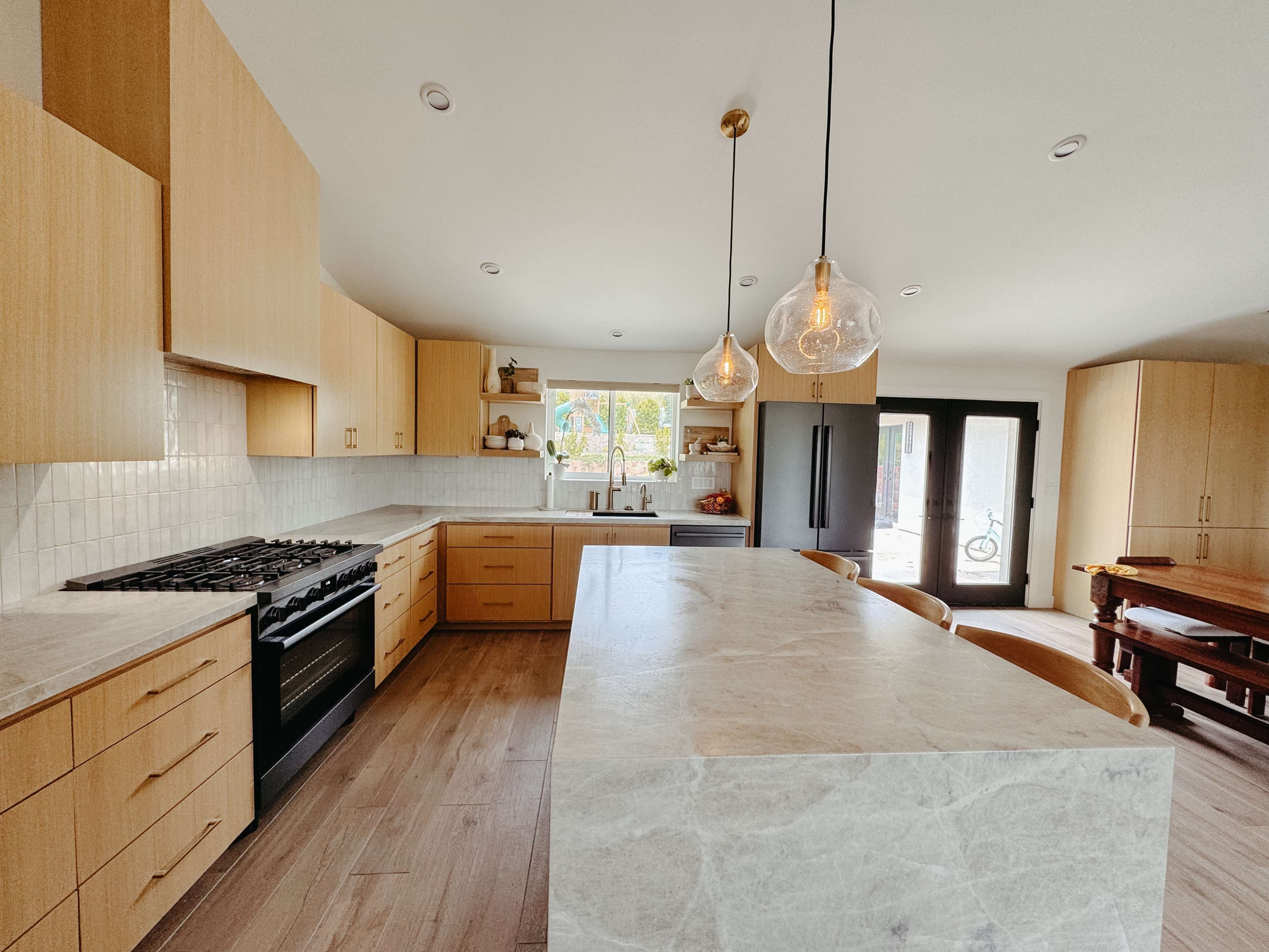A modern kitchen features wooden cabinetry, a large marble island, and pendant lighting above the dining area.