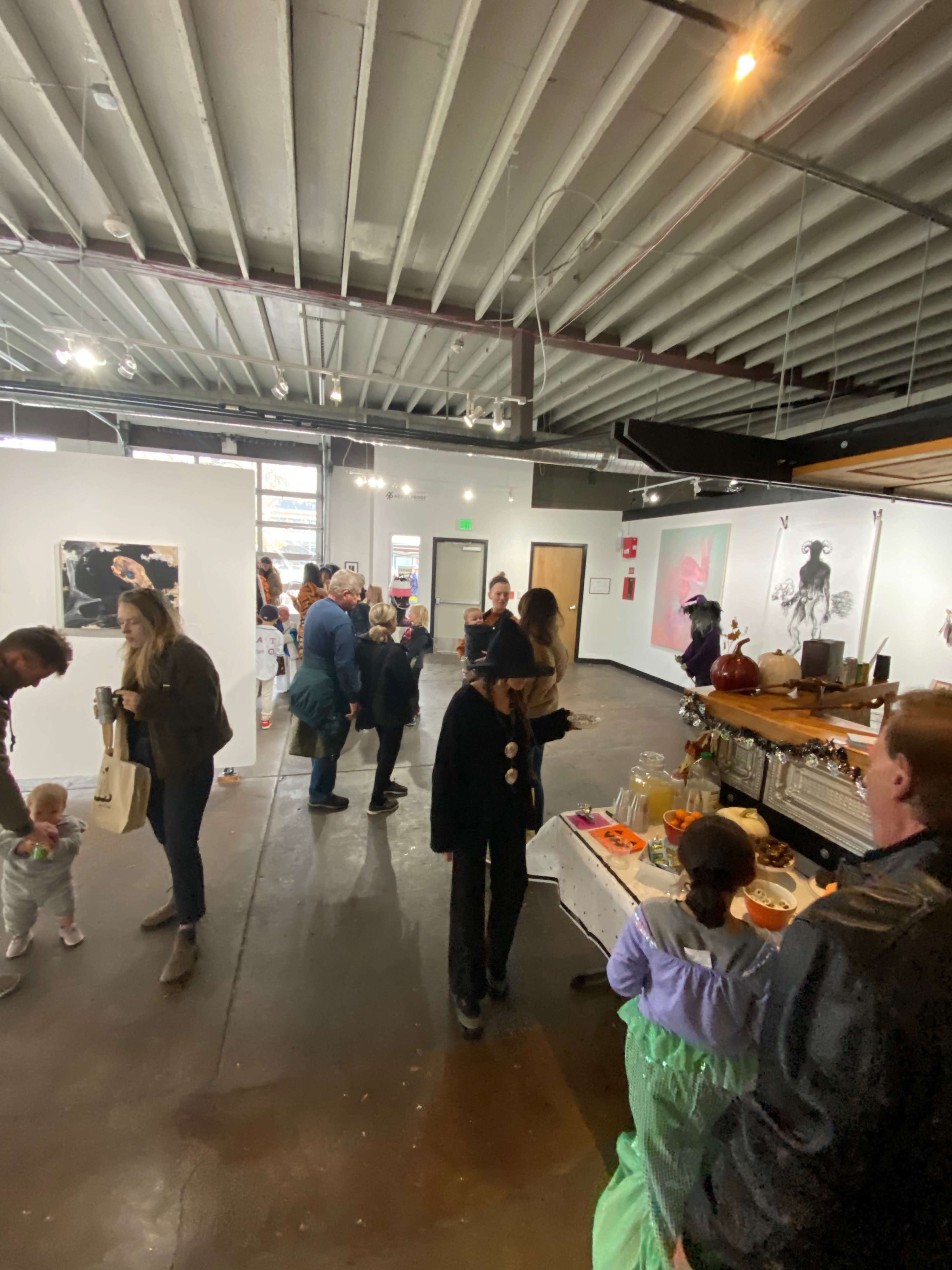 A crowd of people mingle inside an art gallery featuring white walls and various exhibits, with a refreshments table in the foreground.