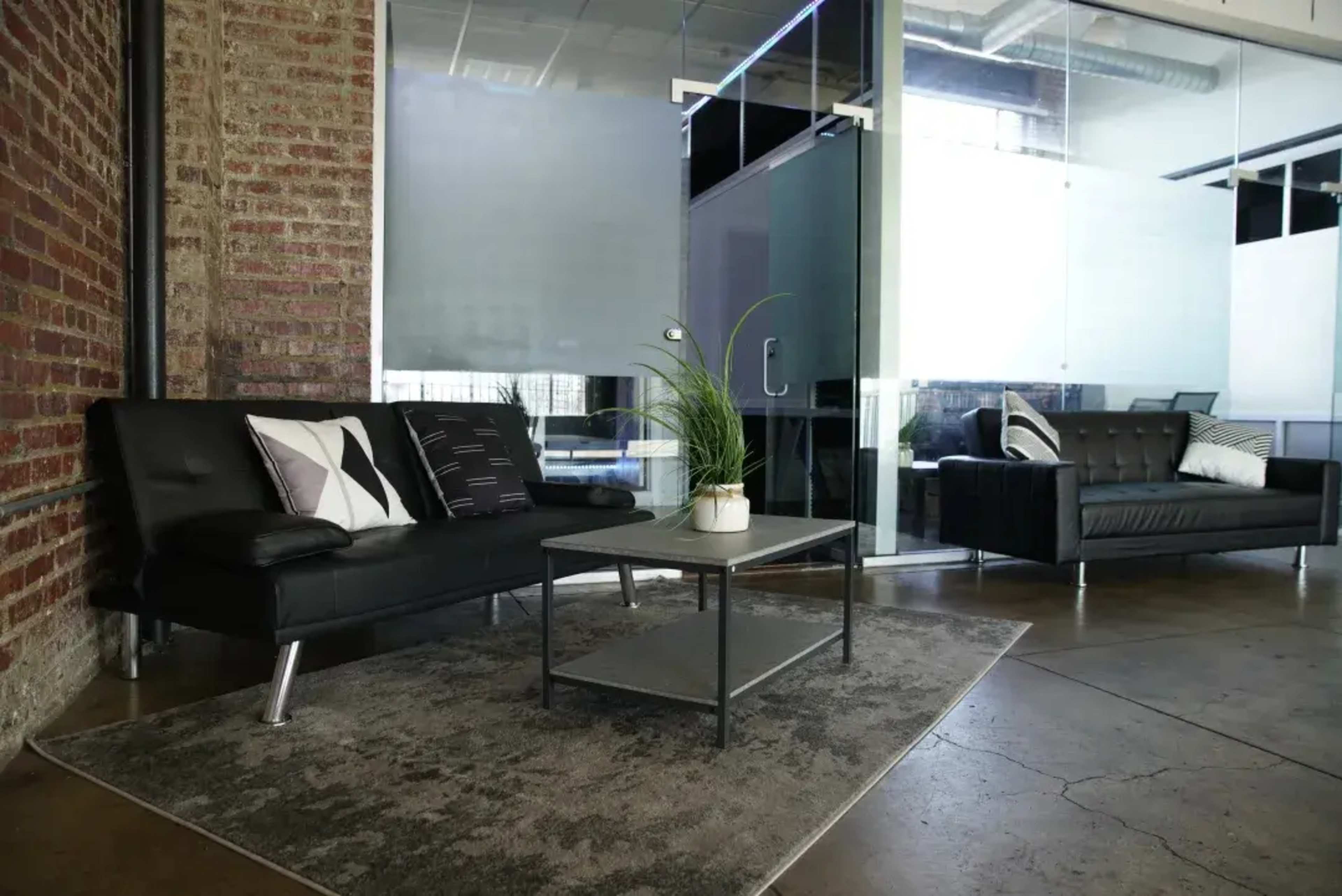 The image shows a modern office lounge area featuring two black sofas, a coffee table, and a potted plant on a rug, with exposed brick walls and glass partitions in the background.