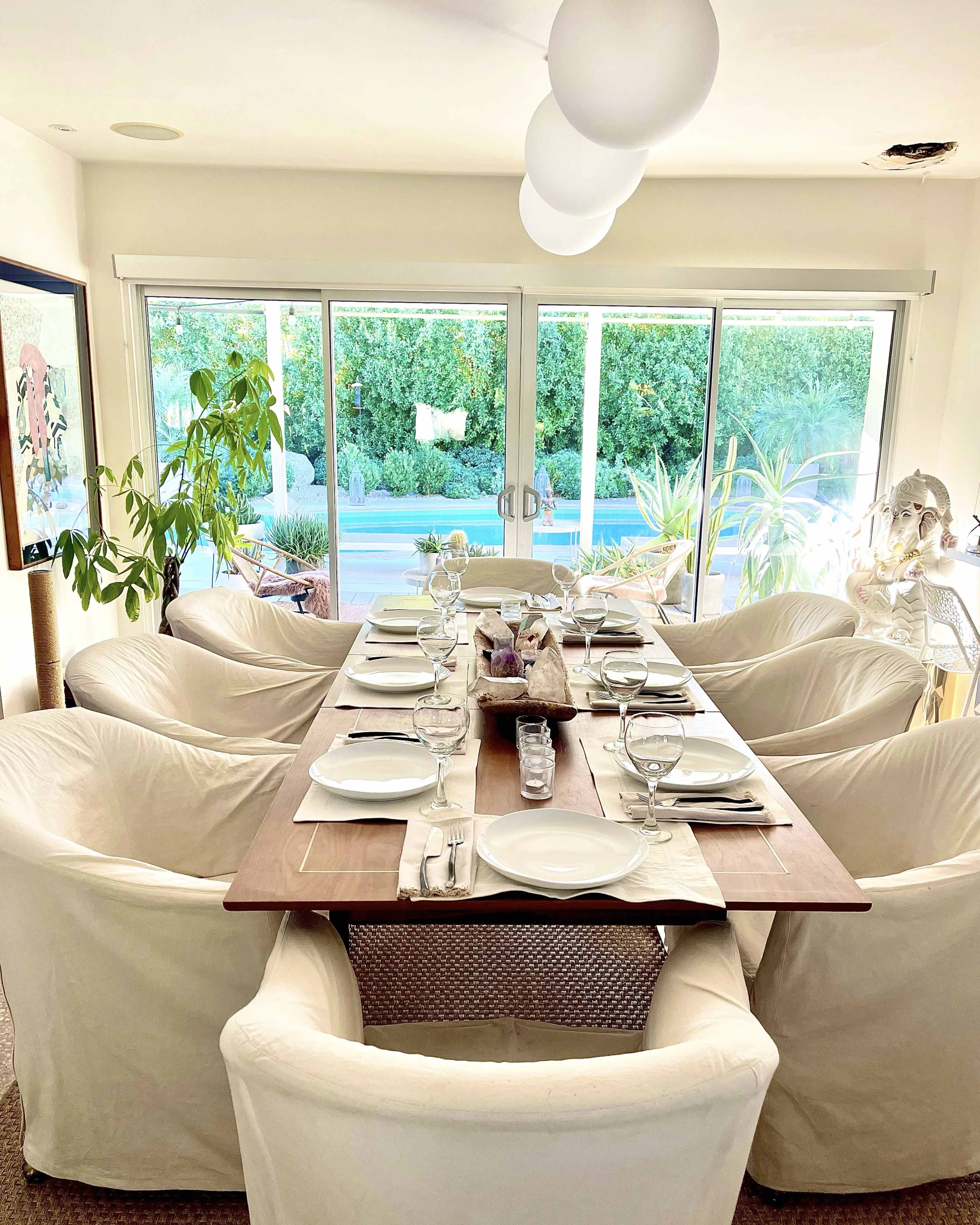 A long wooden dining table is set with white plates and glassware, surrounded by white upholstered chairs in a bright room with large windows overlooking greenery.