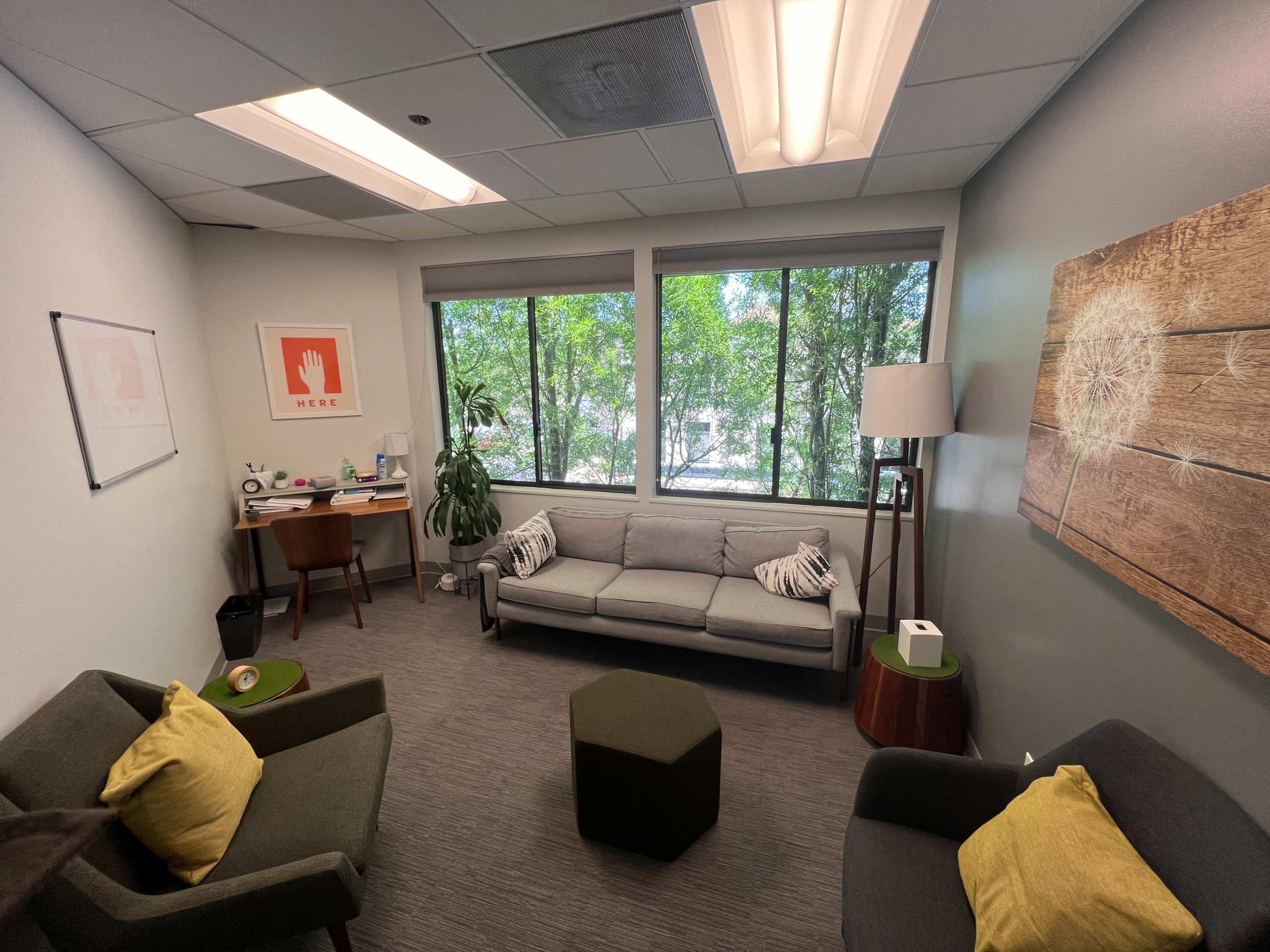 A cozy office space features a gray couch, two green chairs, a wooden desk, and large windows with views of trees outside.