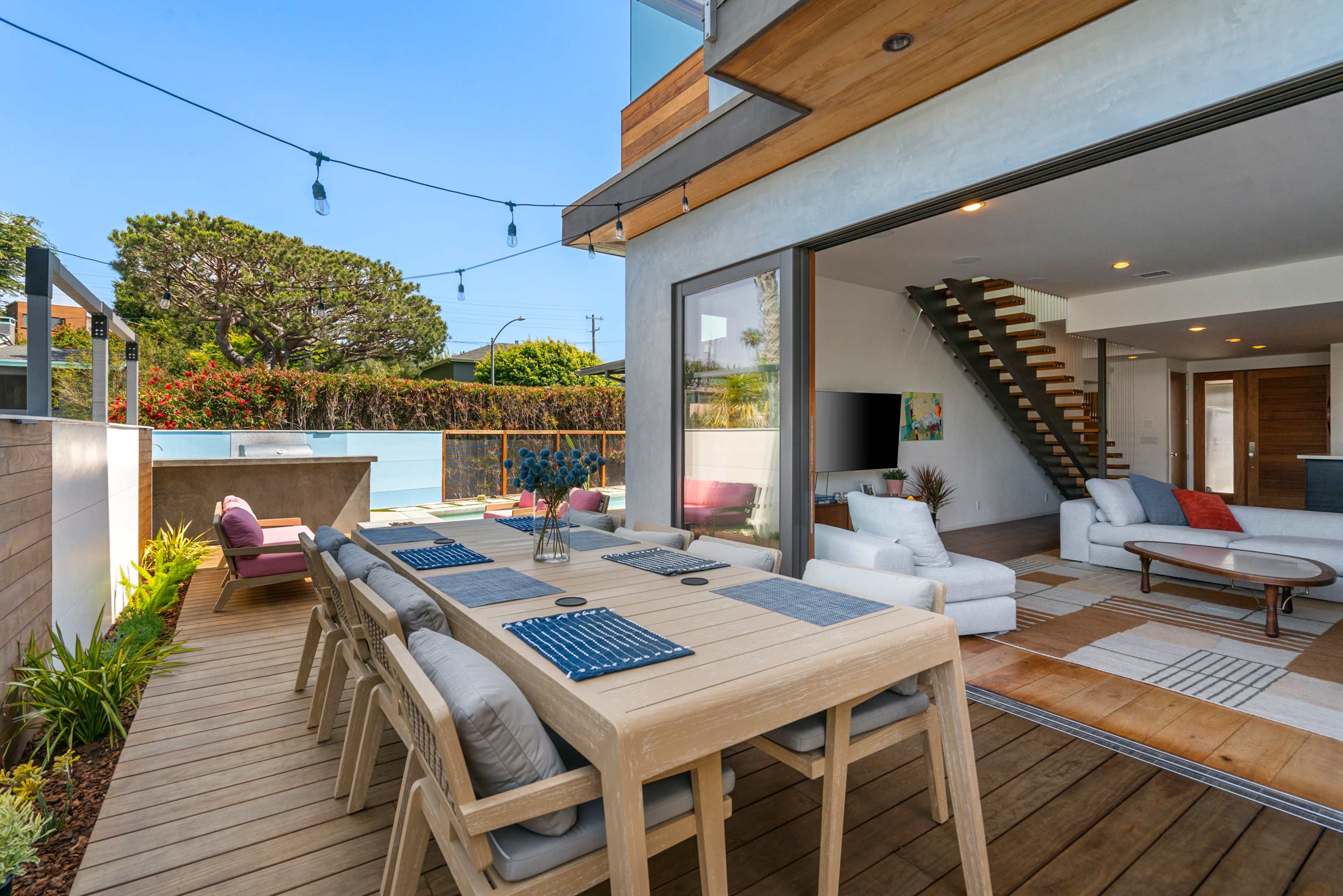 A modern outdoor dining area with a wooden table and chairs, leading to an open living space featuring a staircase and large windows.
