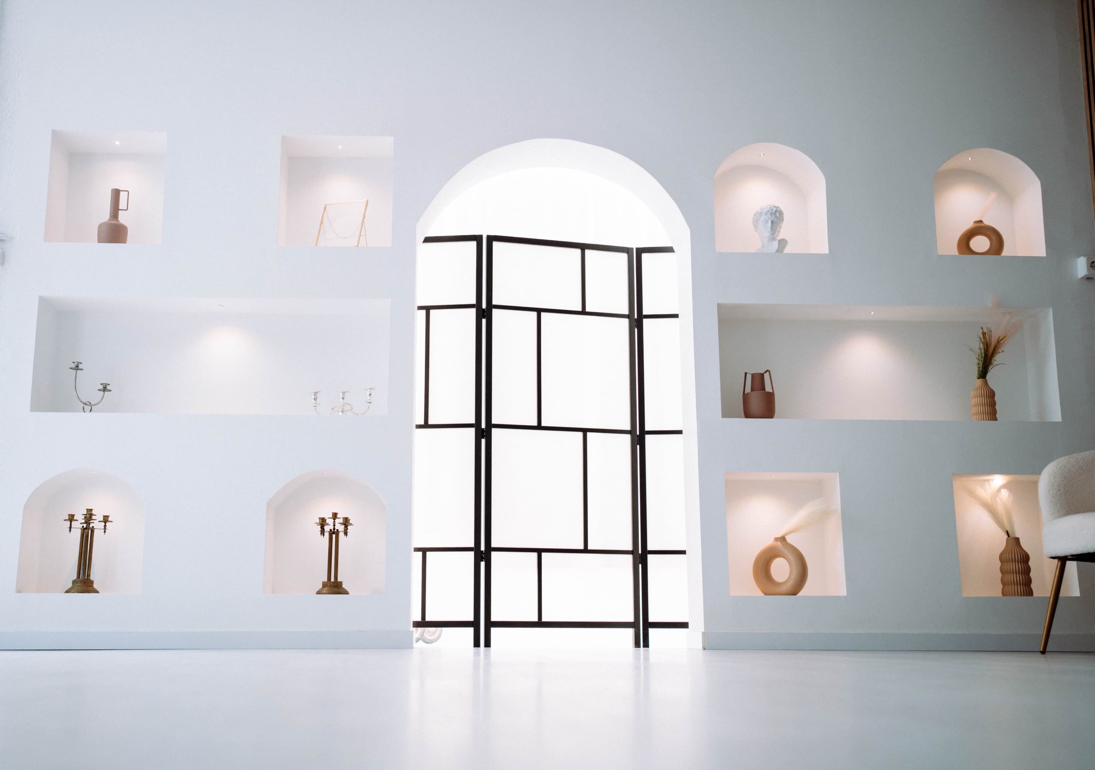 A minimalist room features a black open frame door with a white archway, surrounded by built-in shelves displaying decorative objects.