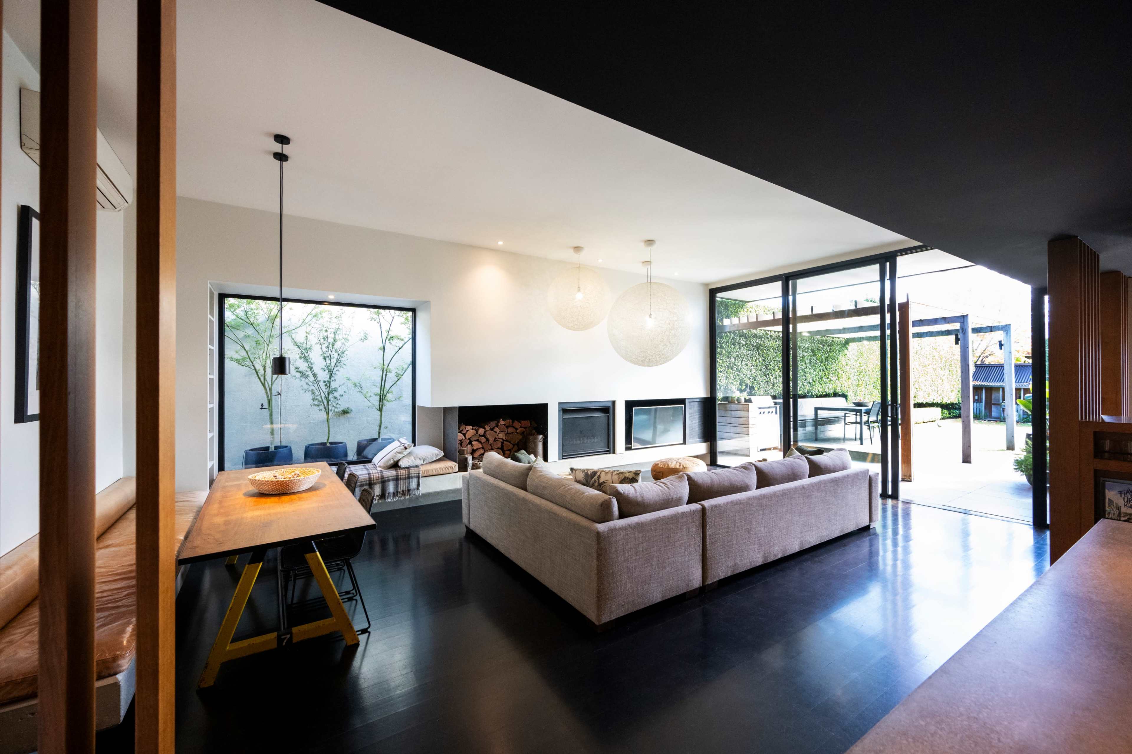 The image shows a modern living room featuring large windows, a comfortable sofa, a dining table, and a fireplace, with an outdoor patio visible through the glass doors.