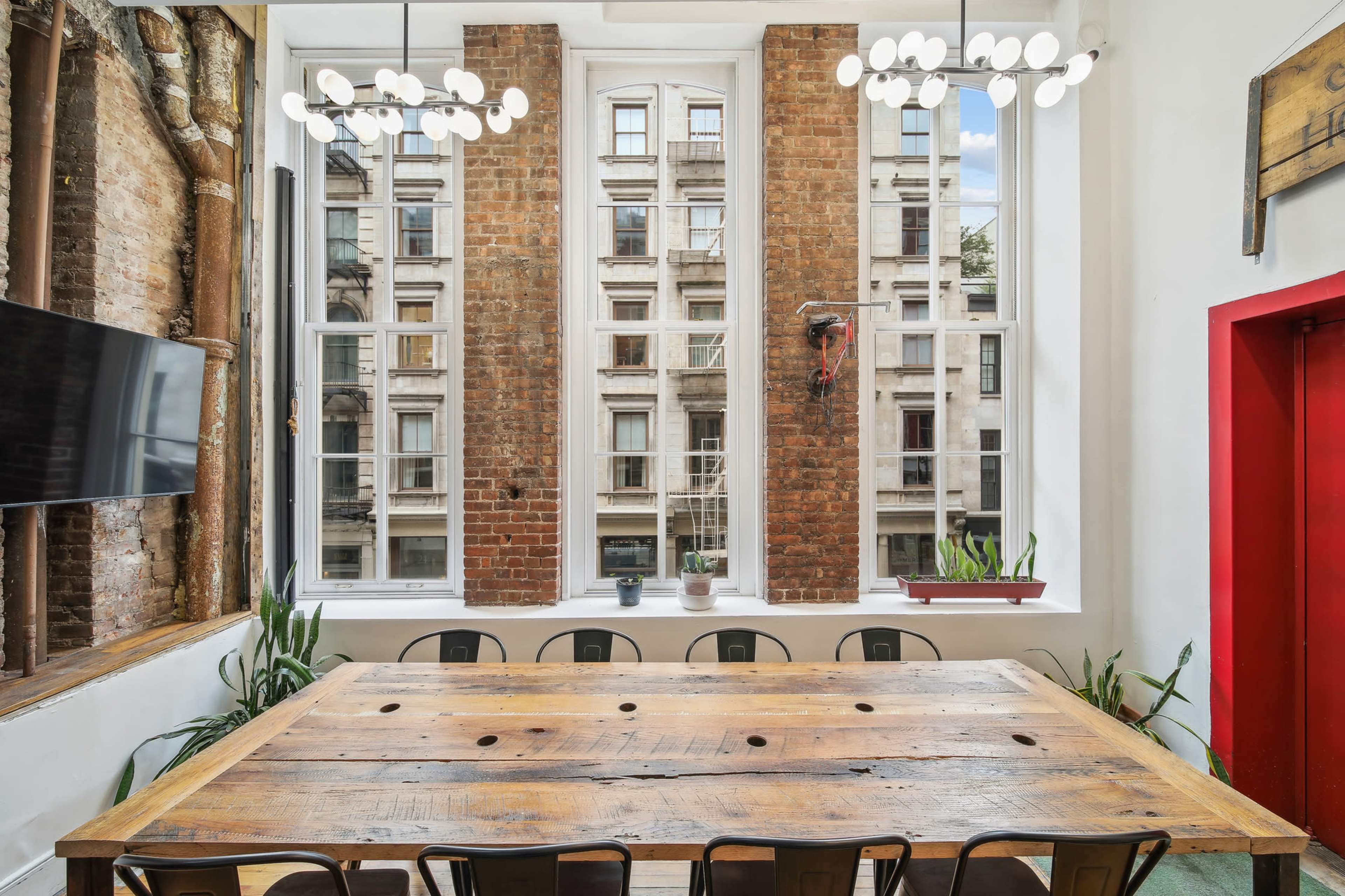 A long wooden table with several chairs sits in a bright room featuring large windows, exposed brick walls, and modern light fixtures.