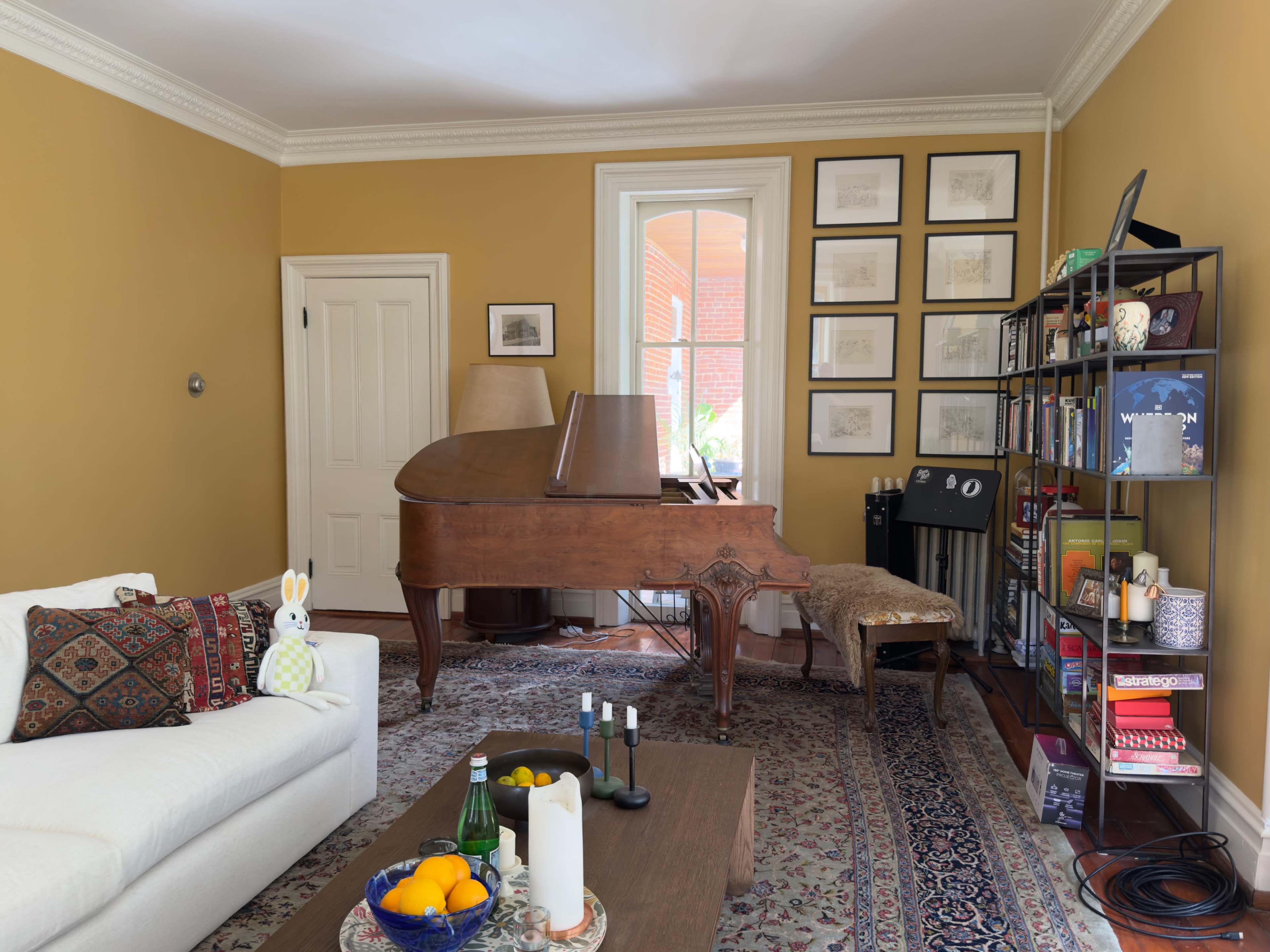 A living room features a grand piano, a shelf filled with books and decor, a light-colored sofa with decorative pillows, and a coffee table with fruits and candles.
