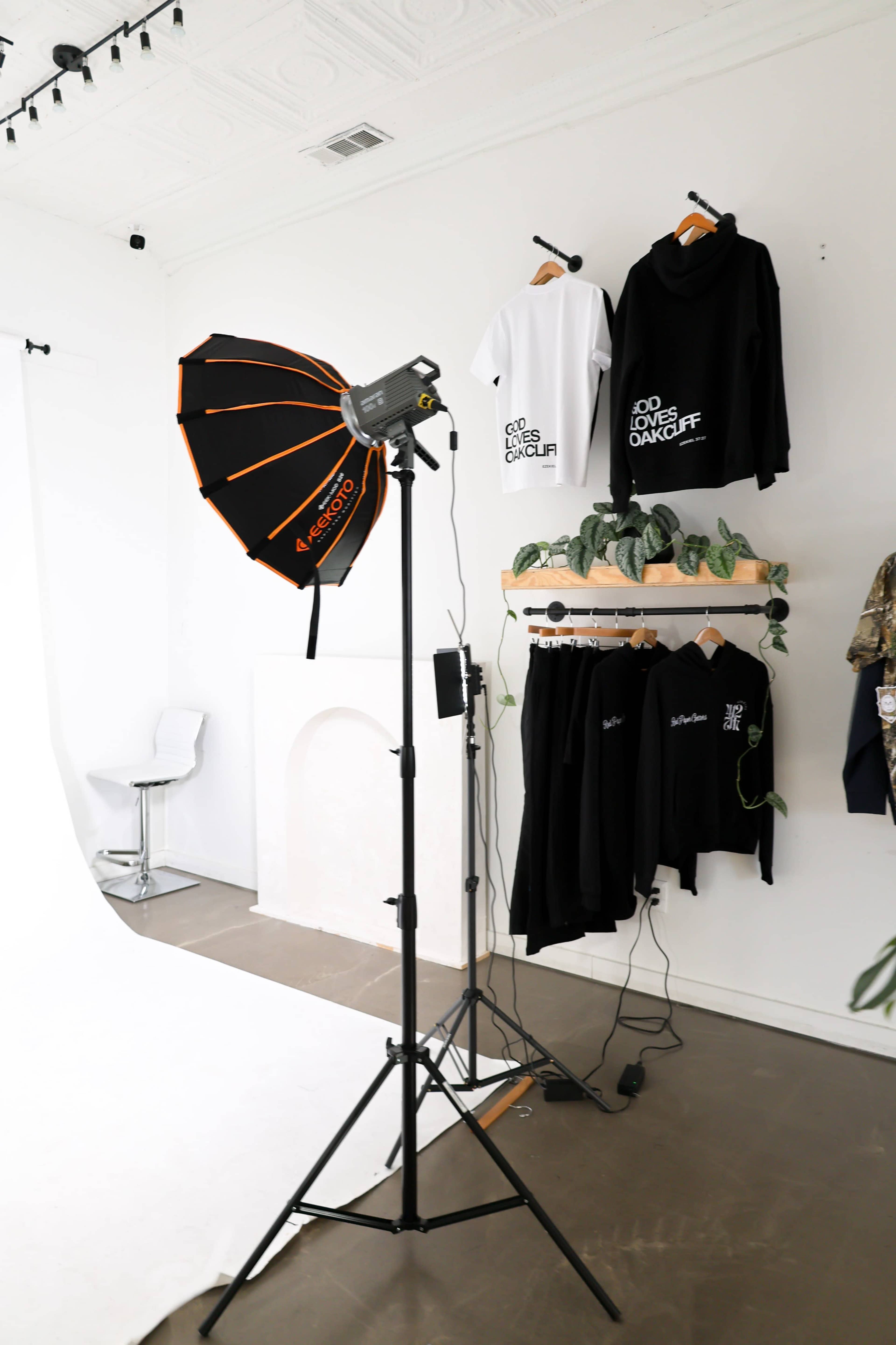 A photography setup features a softbox light on a stand, clothing displays on a wall, and a white backdrop in a studio environment.