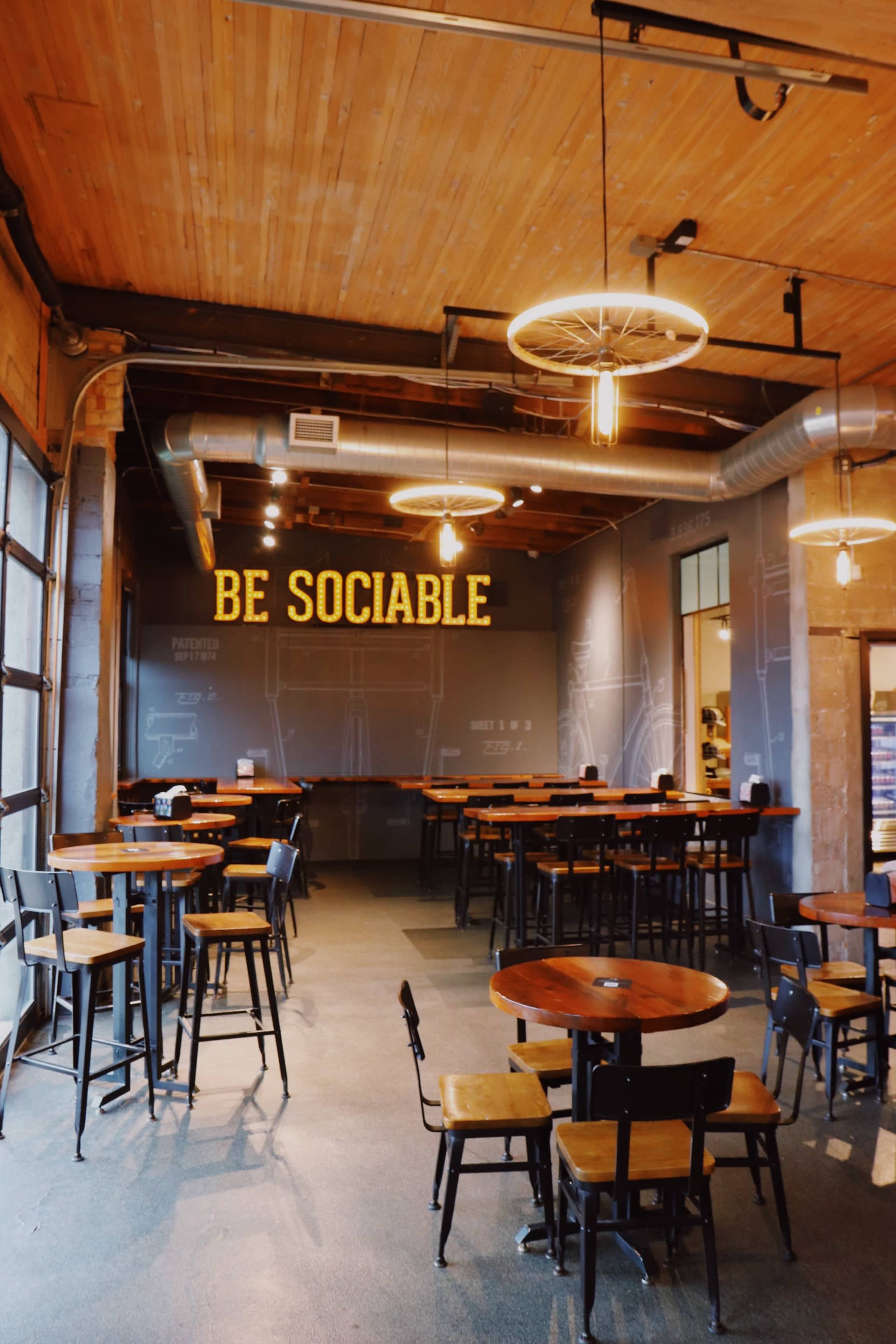 The interior of a café features wooden tables and chairs, with a large sign reading "BE SOCIABLE" on the wall.