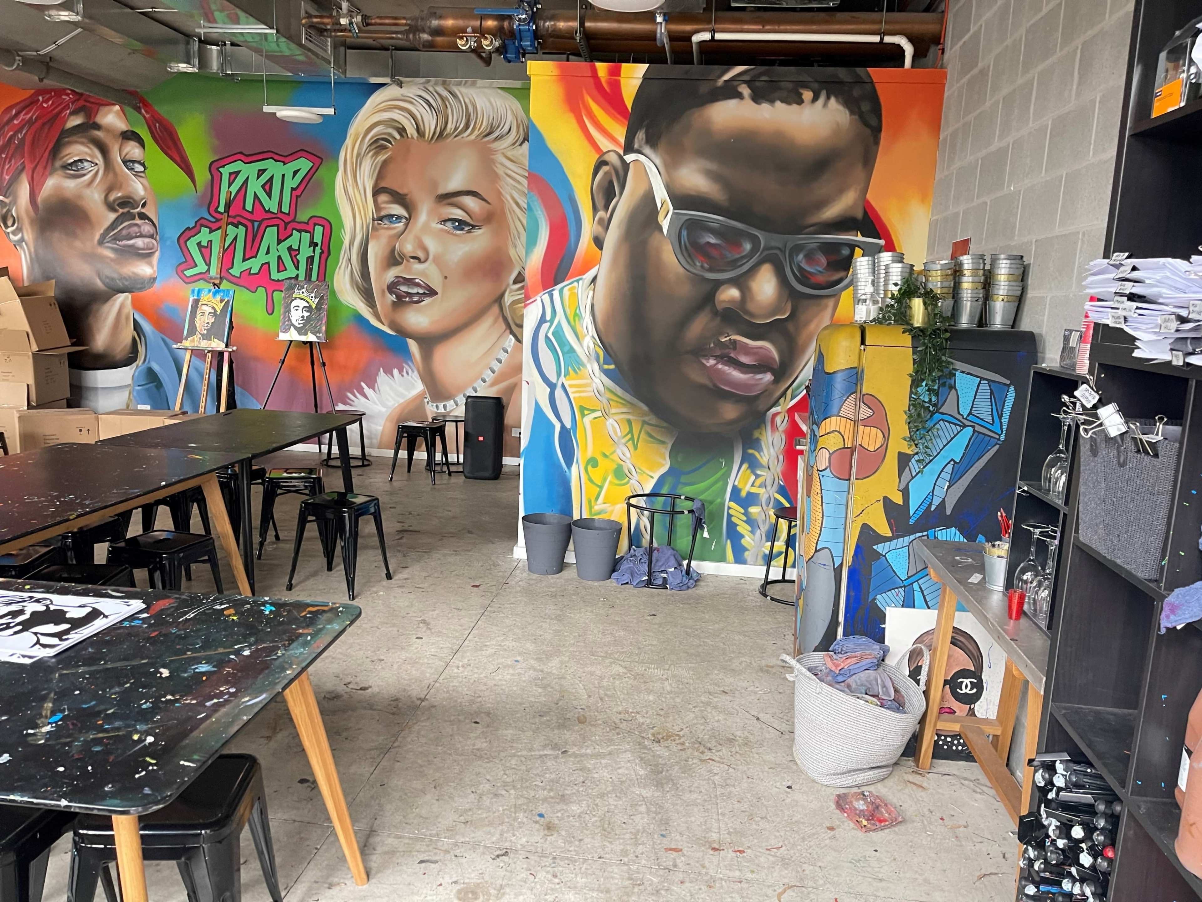 A vibrant art studio featuring large murals of prominent figures on the walls, along with black tables and various art supplies scattered throughout the space.