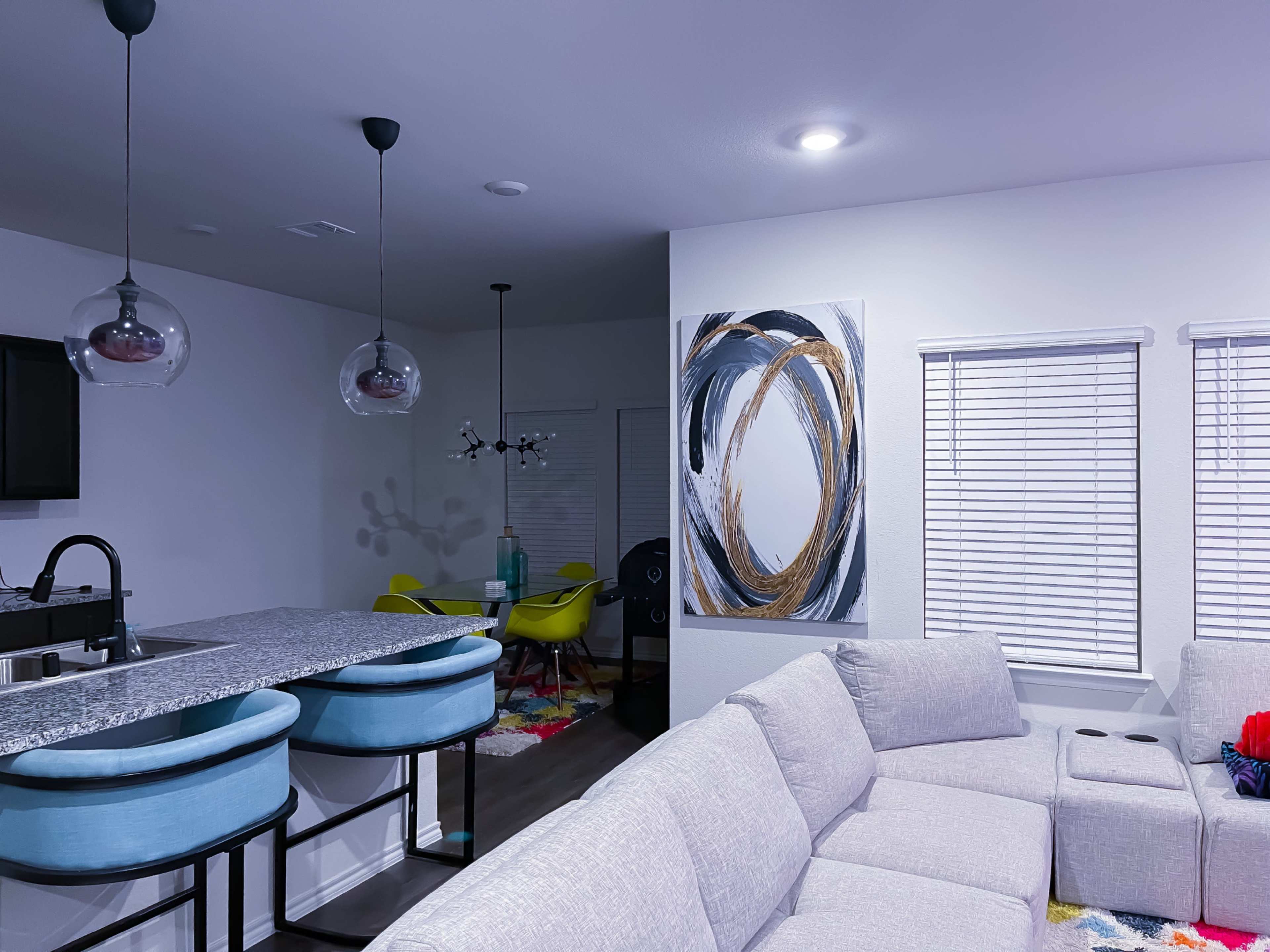 A modern living space features a light-colored sectional sofa, a kitchen island with blue bar stools, and abstract wall art above, with white blinds on the windows.