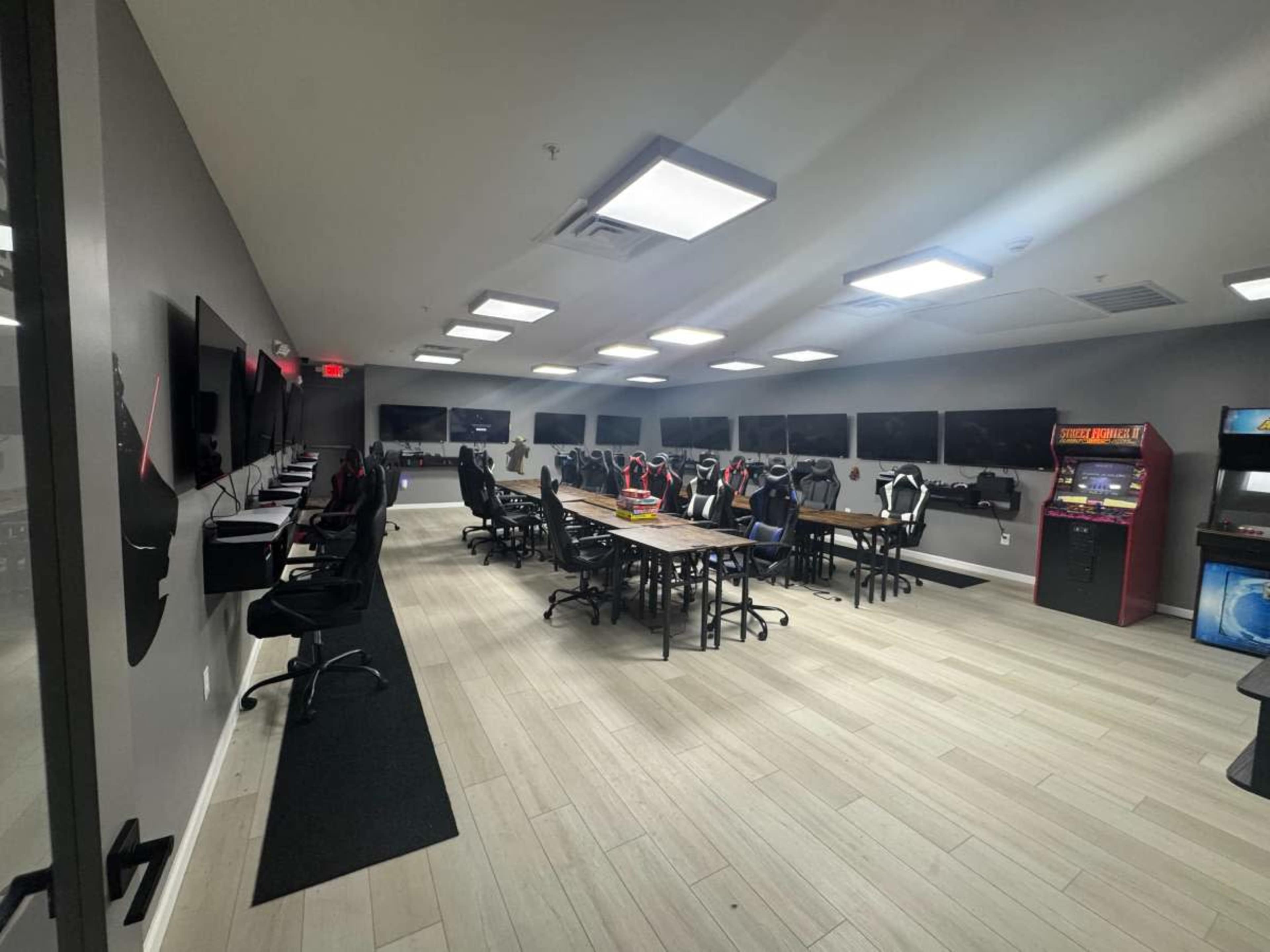 The image shows a modern gaming room featuring multiple computer stations on one side, a long conference-style table in the center, and an arcade machine in the corner.