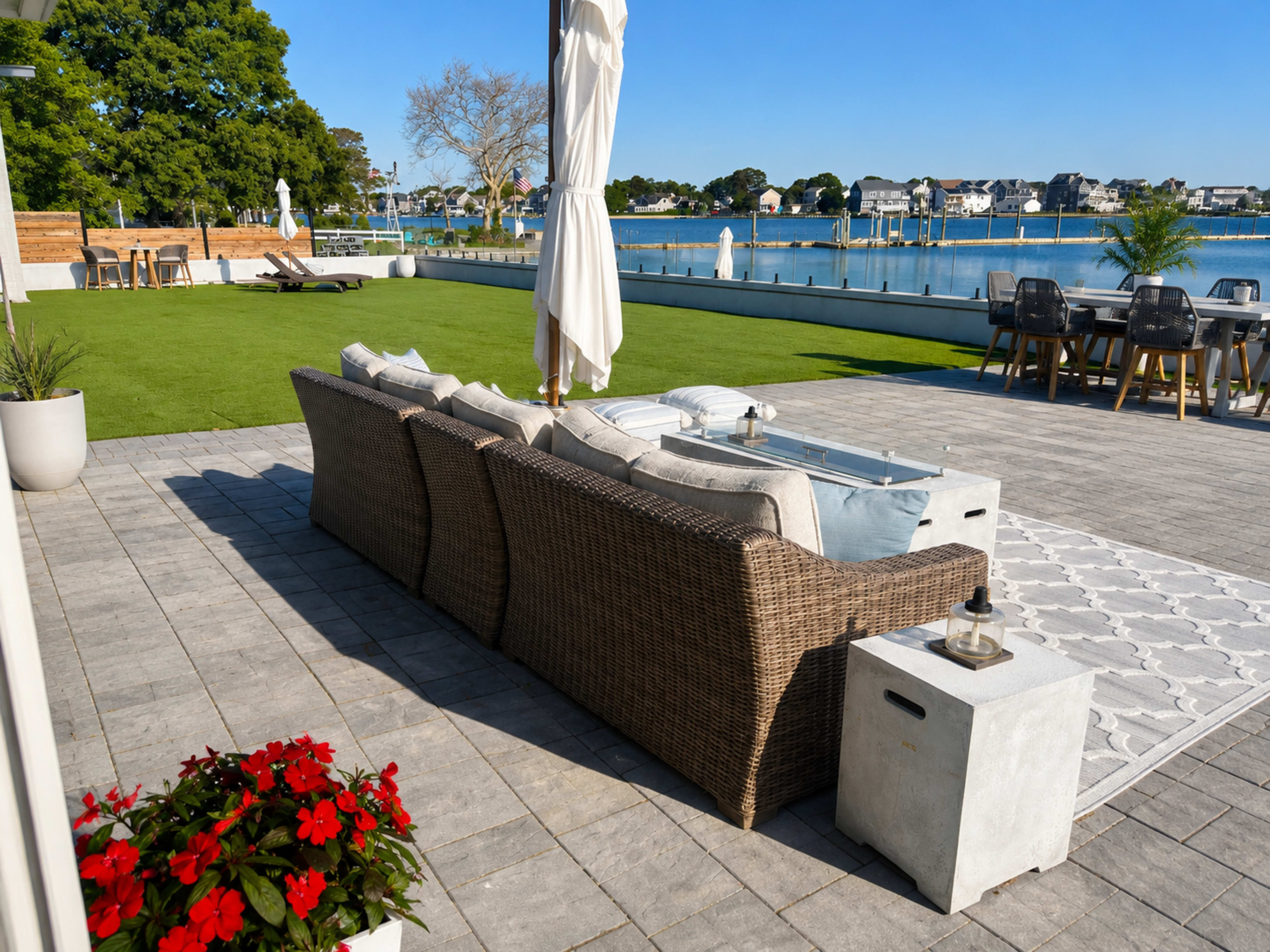 A patio with a woven sofa, a small table, and a well-manicured lawn overlooks a calm waterway with houses in the background.