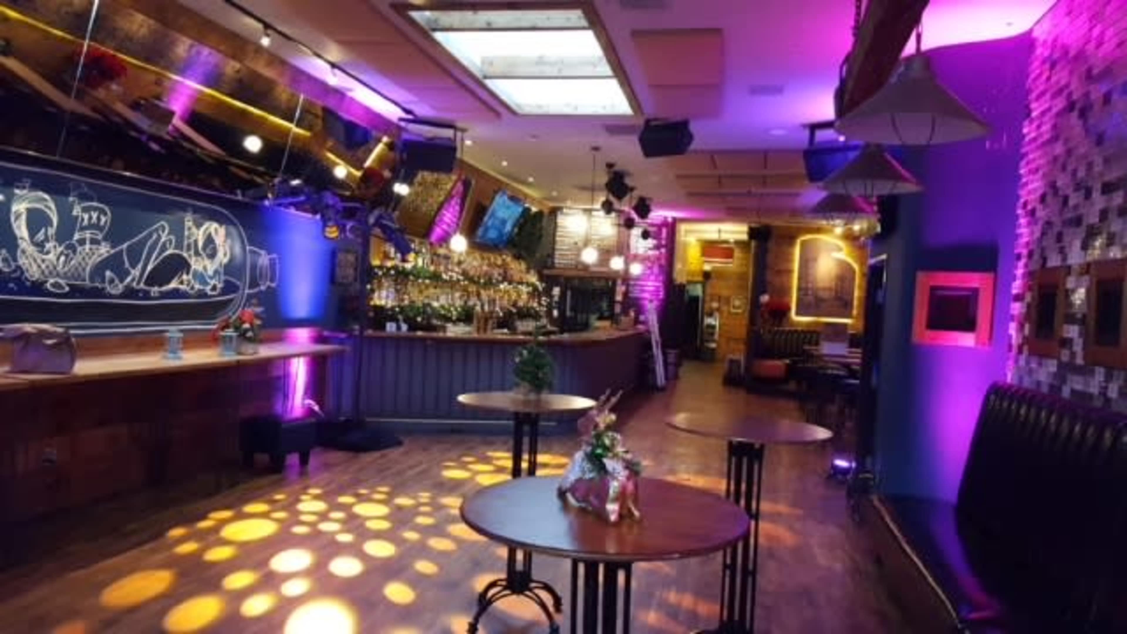 A contemporary bar interior featuring tables, a long bar with bottles, and purple lighting accents.